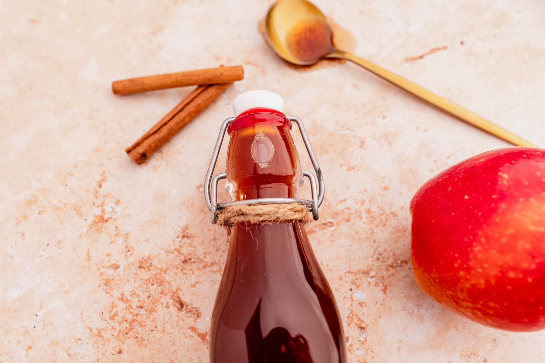 A sealed glass bottle filled with brown liquid, a red apple, cinnamon sticks, and a spoon with Starbucks apple crisp syrup residue rest on a textured beige surface.