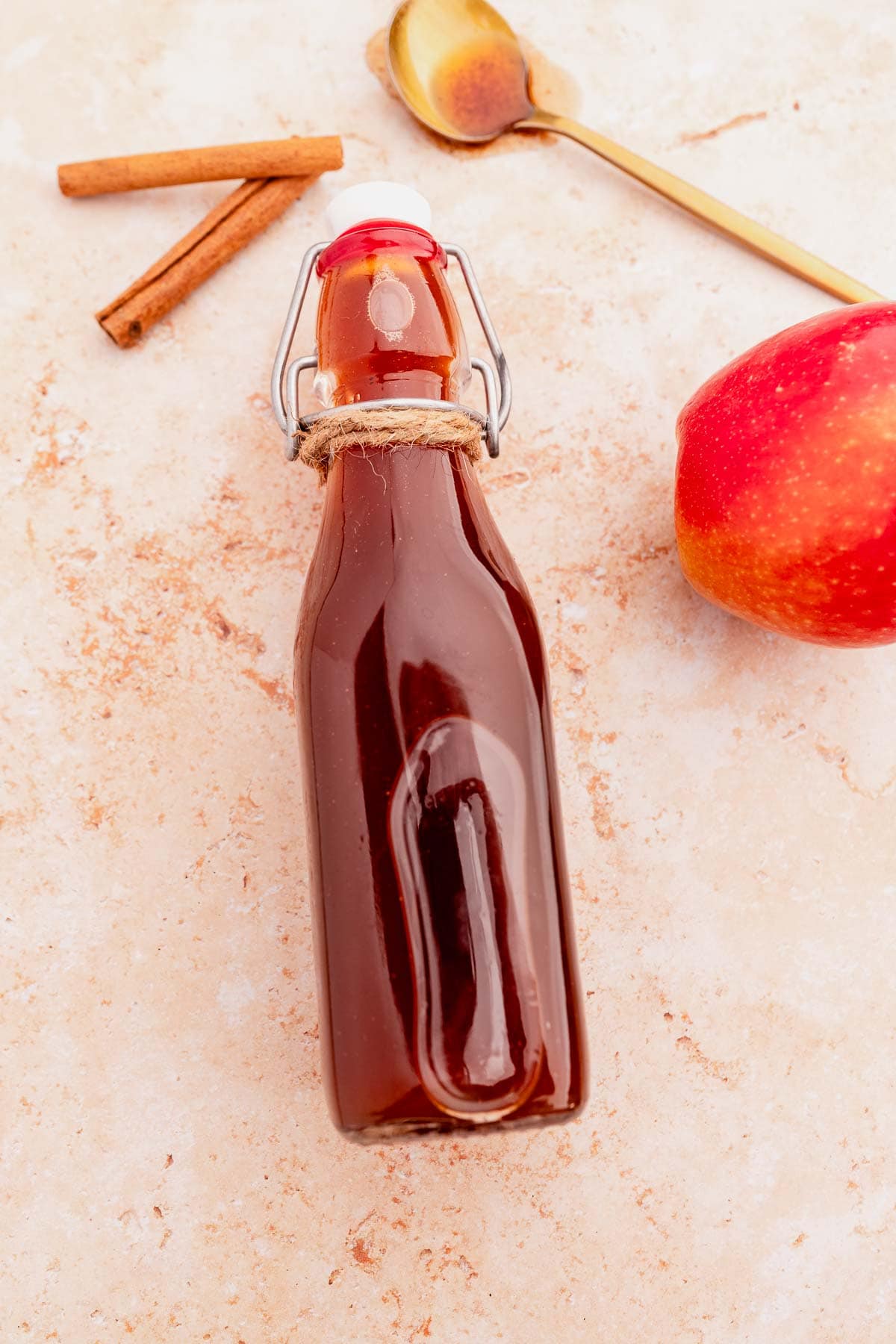 A glass bottle with dark liquid, surrounded by cinnamon sticks and a metal spoon, sits next to a red apple and a hint of Starbucks apple crisp syrup on a light textured surface.