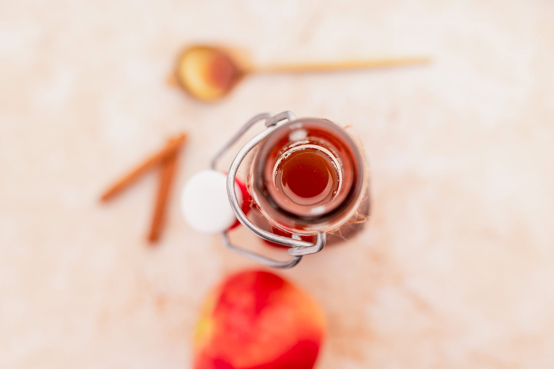 Top view of an open glass bottle filled with amber liquid, reminiscent of Starbucks apple crisp syrup, with a blurred red apple, cinnamon sticks, and a wooden spoon in the background.