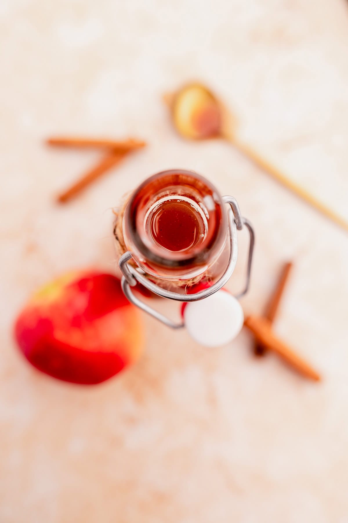 A bottle of apple cider vinegar is seen from above, surrounded by a red apple, cinnamon sticks, and a wooden spoon on a beige surface, evoking the warm flavors reminiscent of Starbucks apple crisp syrup.
