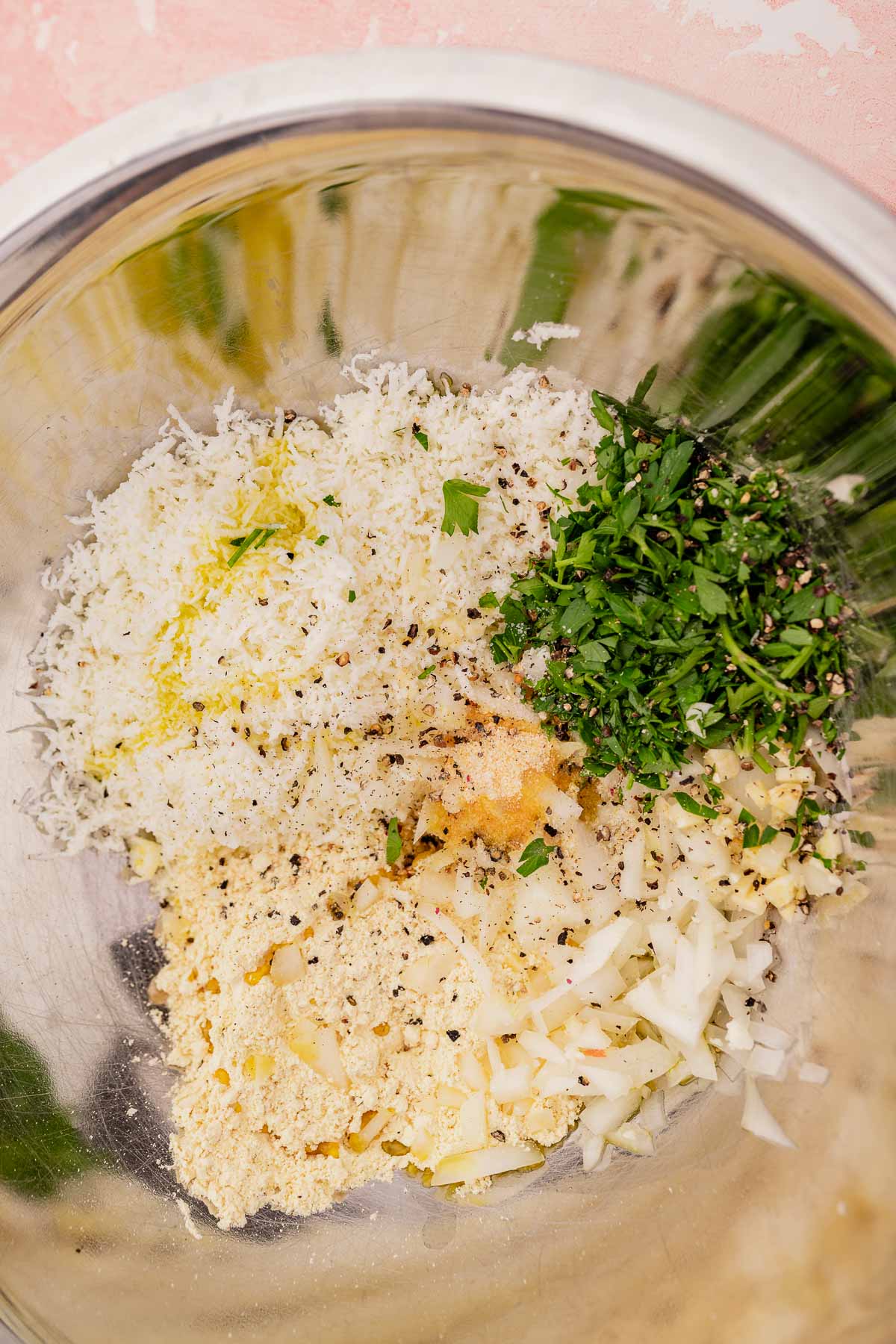 A metal bowl containing shredded cheese, chopped herbs, diced onions, spices, and flour mixed together forms the perfect base for mouthwatering halloumi fritters.