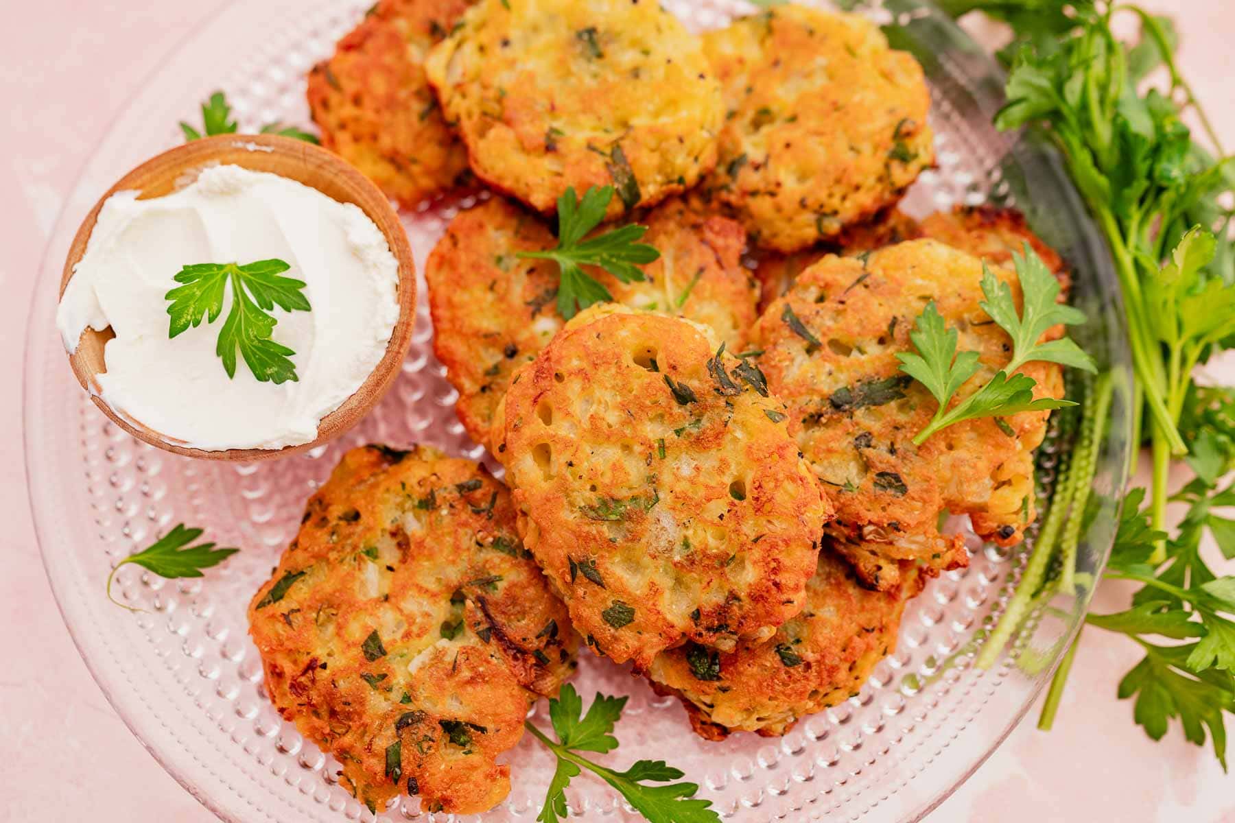 A plate of herb-flecked halloumi fritters garnished with parsley. Accompanied by a small bowl of white dipping sauce and fresh parsley sprigs on a clear glass plate.