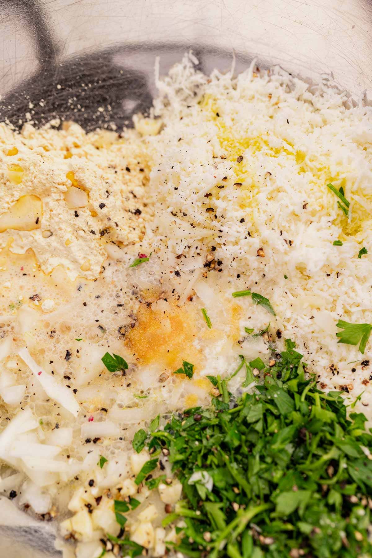 Close-up of ingredients in a bowl, ready for halloumi fritters: grated cheese, chopped parsley, diced onions, ground pepper, and a yellowish powder await mixing.