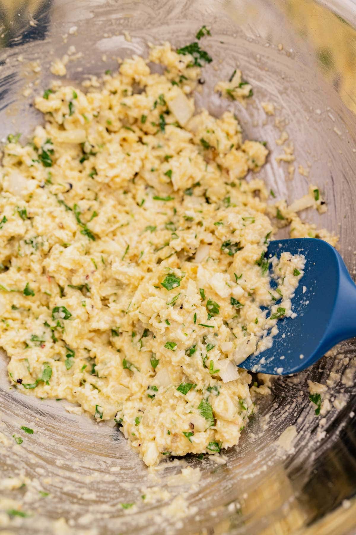 A bowl of mixed eggs, herbs, spices, and grated halloumi fritters with a blue spatula.