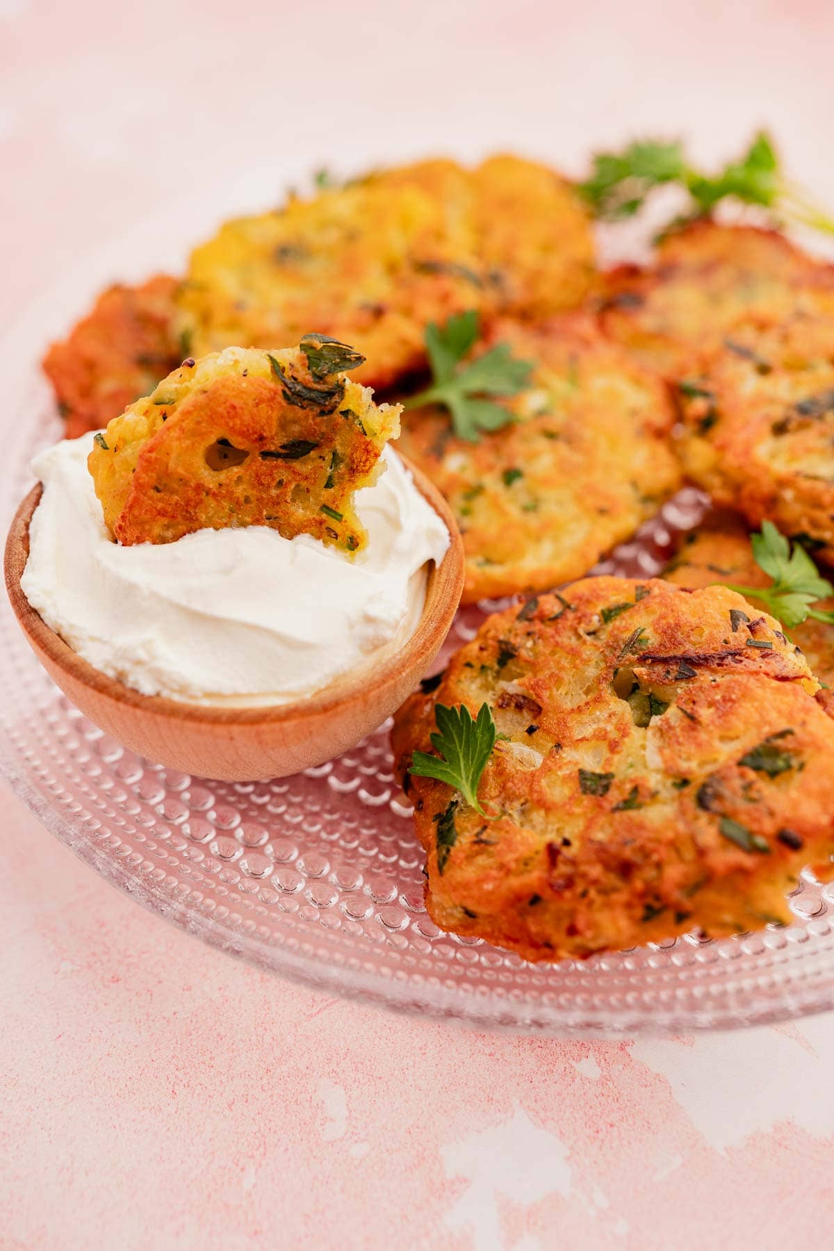 A clear plate showcases golden-brown vegetable and halloumi fritters, garnished with herbs and accompanied by a small wooden bowl of creamy dip, all elegantly set on a light pink surface.