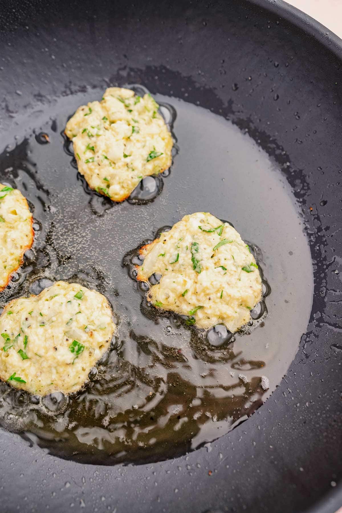 Three halloumi fritters sizzling in oil in a black pan.