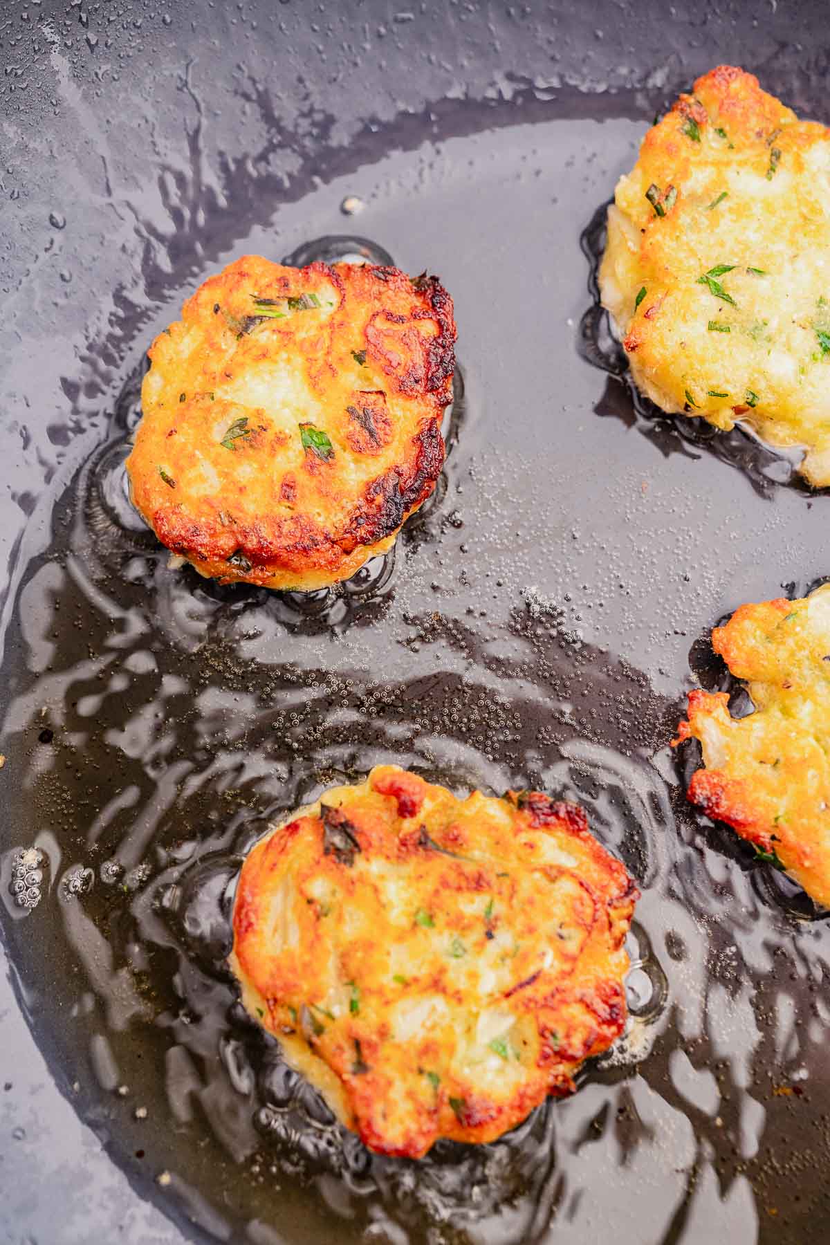Four golden-brown halloumi fritters sizzling in oil in a black frying pan.