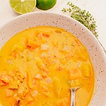 A bowl of Jamaican pumpkin soup with chunks of vegetables and a spoon. Lime halves and fresh herbs are in the background.