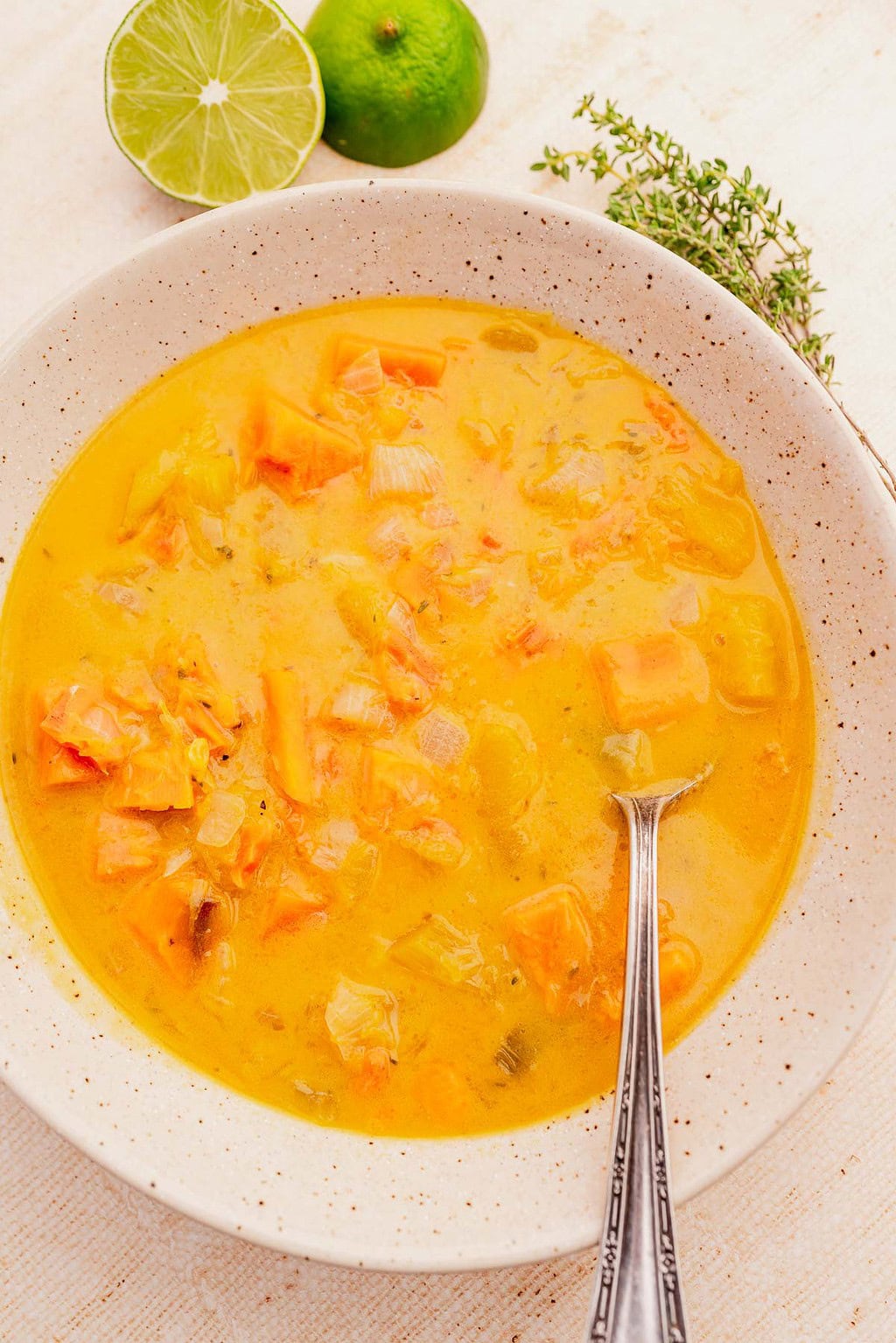 Bowl of orange-yellow Jamaican pumpkin soup with chunks of vegetables, a spoon, and a lime and herbs in the background on a light surface.