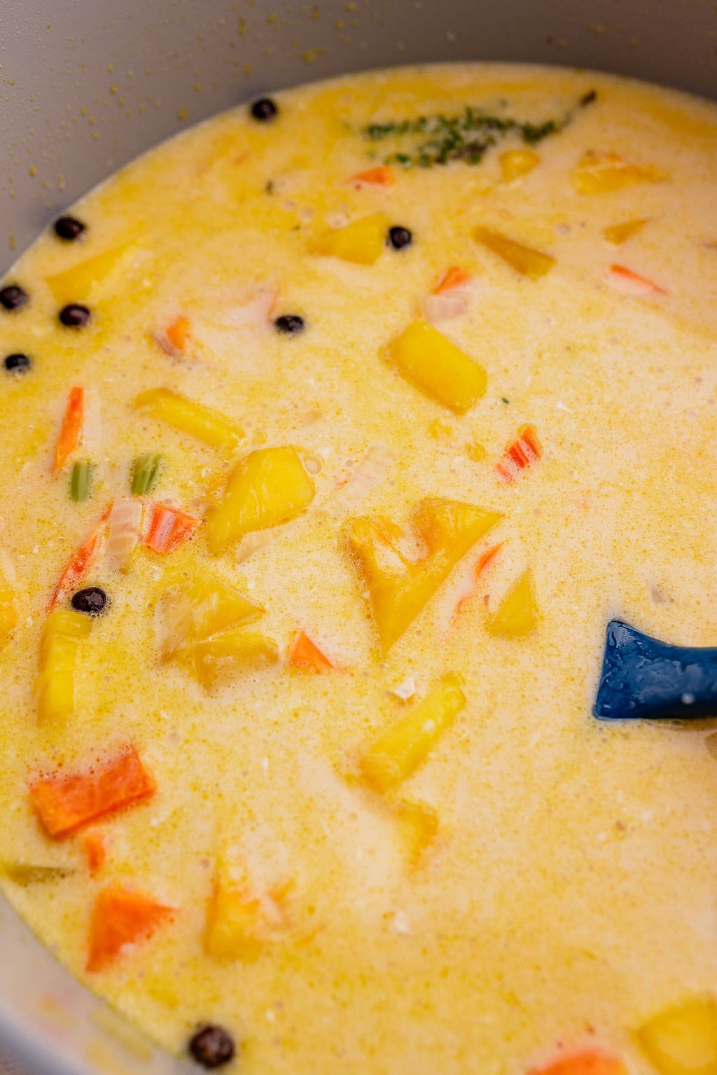 A close-up of a creamy jamaican pumpkin soup with chunks of yellow pineapple, carrots, crab sticks, whole peppercorns, and a blue spatula in a pot.