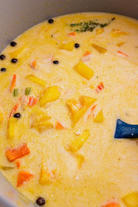 A close-up of a creamy jamaican pumpkin soup with chunks of yellow pineapple, carrots, crab sticks, whole peppercorns, and a blue spatula in a pot.