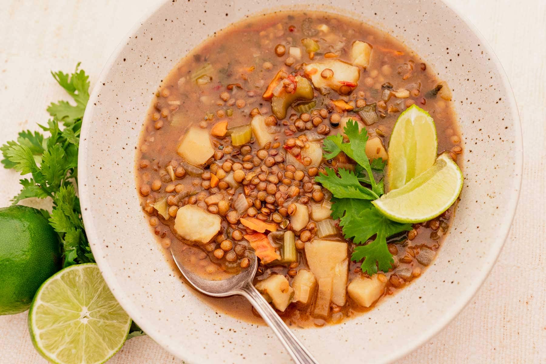 A bowl of lentejas soup with diced vegetables, garnished with lime wedges and cilantro, sits on a beige surface. A spoon rests in the soup, accompanied by fresh lime and cilantro beside the bowl.