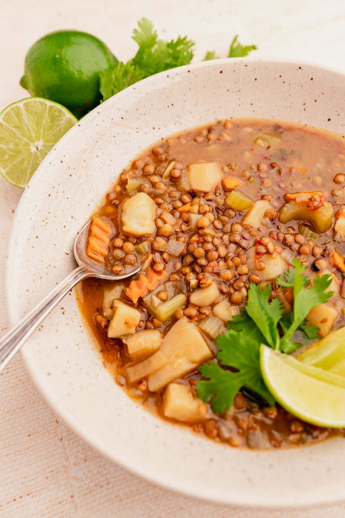 A bowl of lenteja soup with vegetables, garnished with lime wedges and cilantro. A spoon is resting in the bowl, and a whole lime is in the background.