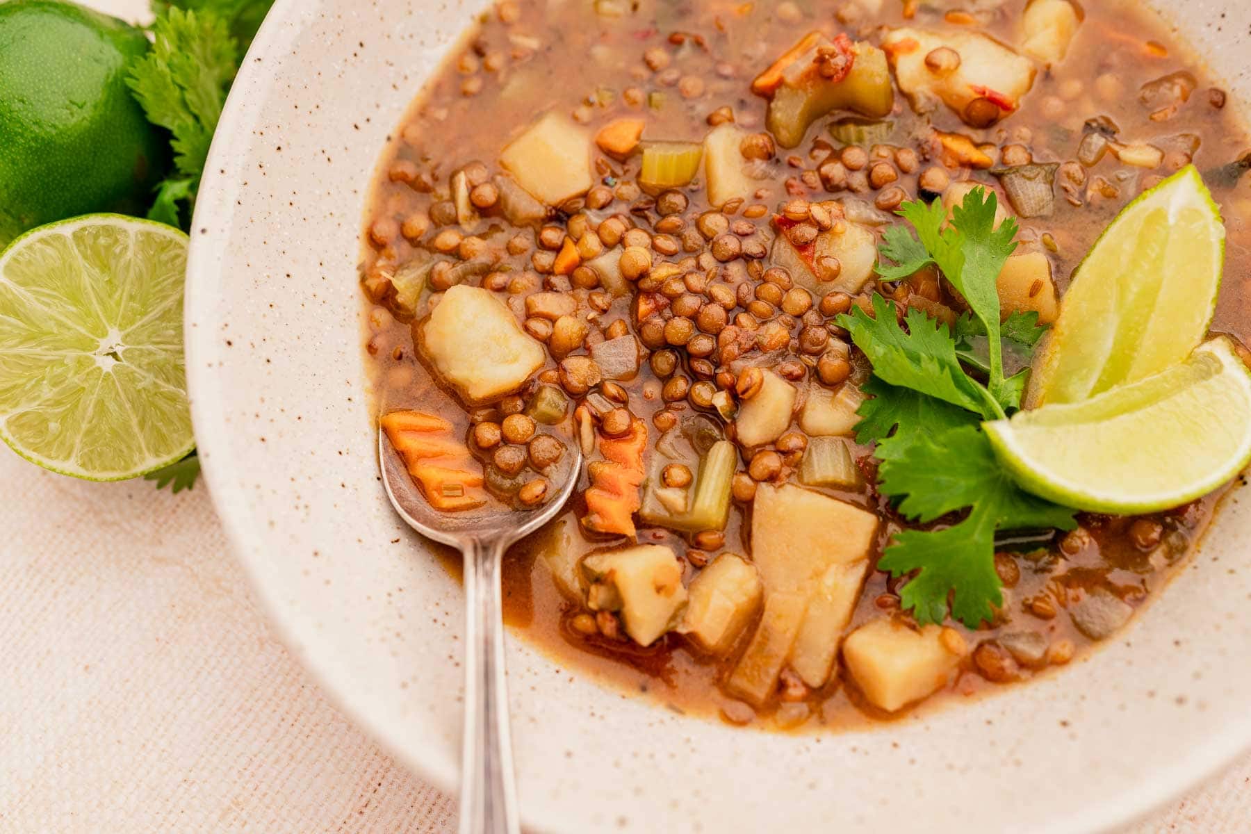 A bowl of vegetable lentil soup, known as lentejas, with diced carrots, potatoes, and celery. Garnished with lime wedges and cilantro.