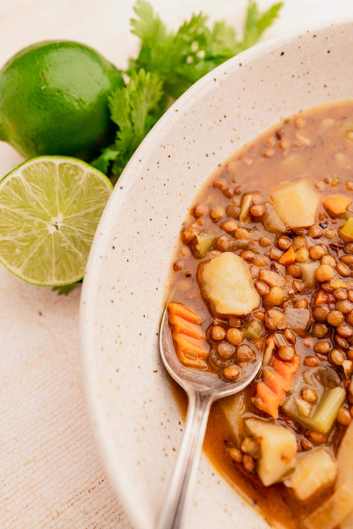 A bowl of lentejas soup with vegetables and a spoon, garnished with lime and cilantro on the side.
