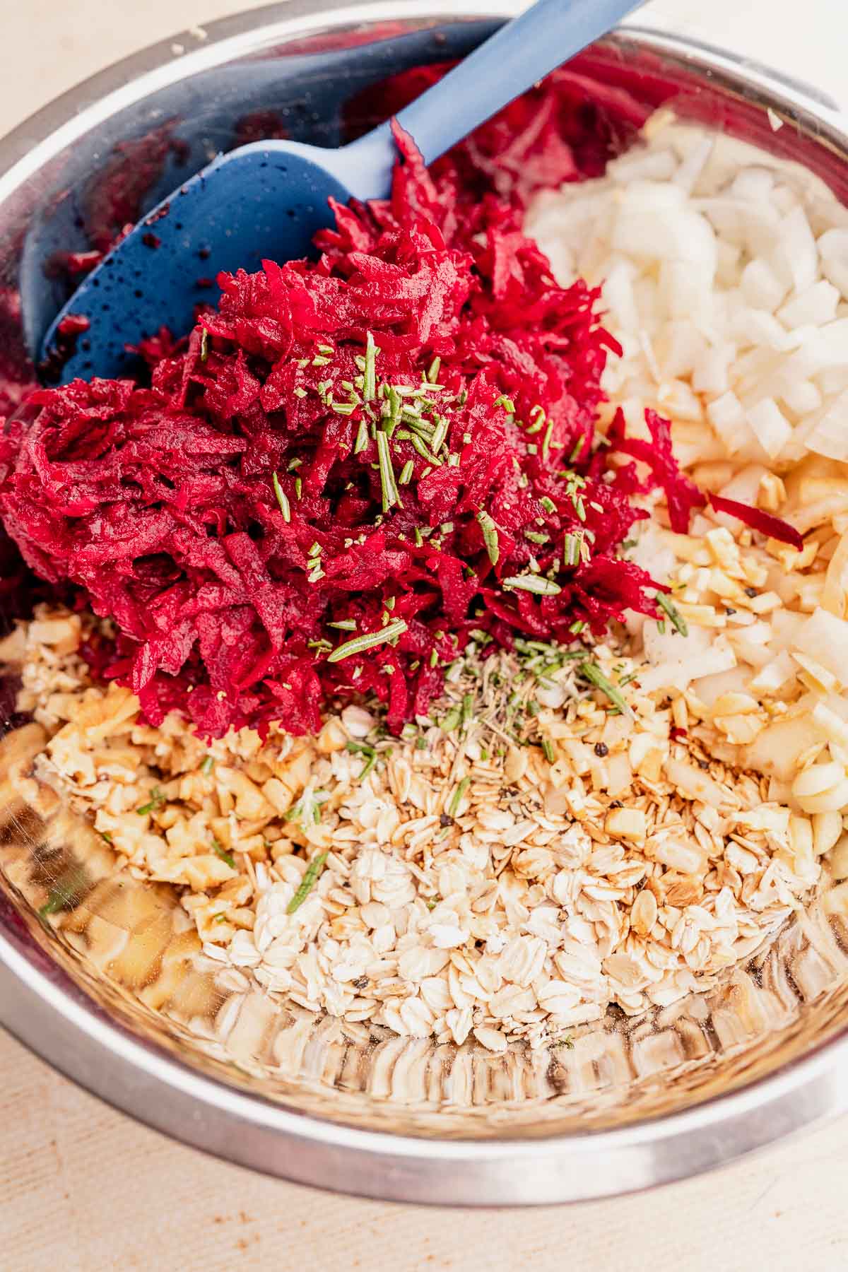 A mixing bowl brimming with grated beetroot, chopped onions, oats, and herbs is expertly combined with a blue spatula to prepare a delicious vegetarian roast.