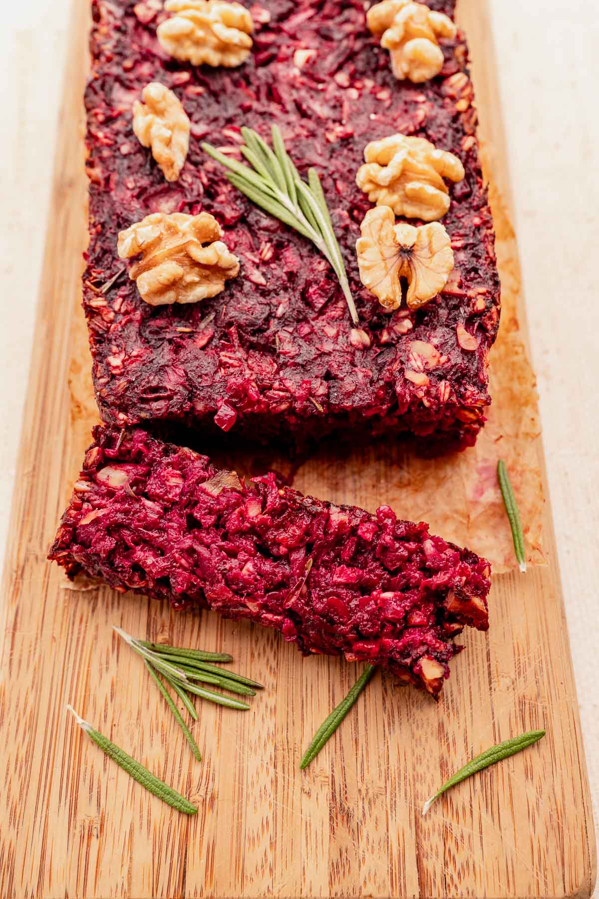 A vegetarian roast loaf on a wooden board, topped with walnuts and rosemary, with one slice partly cut.