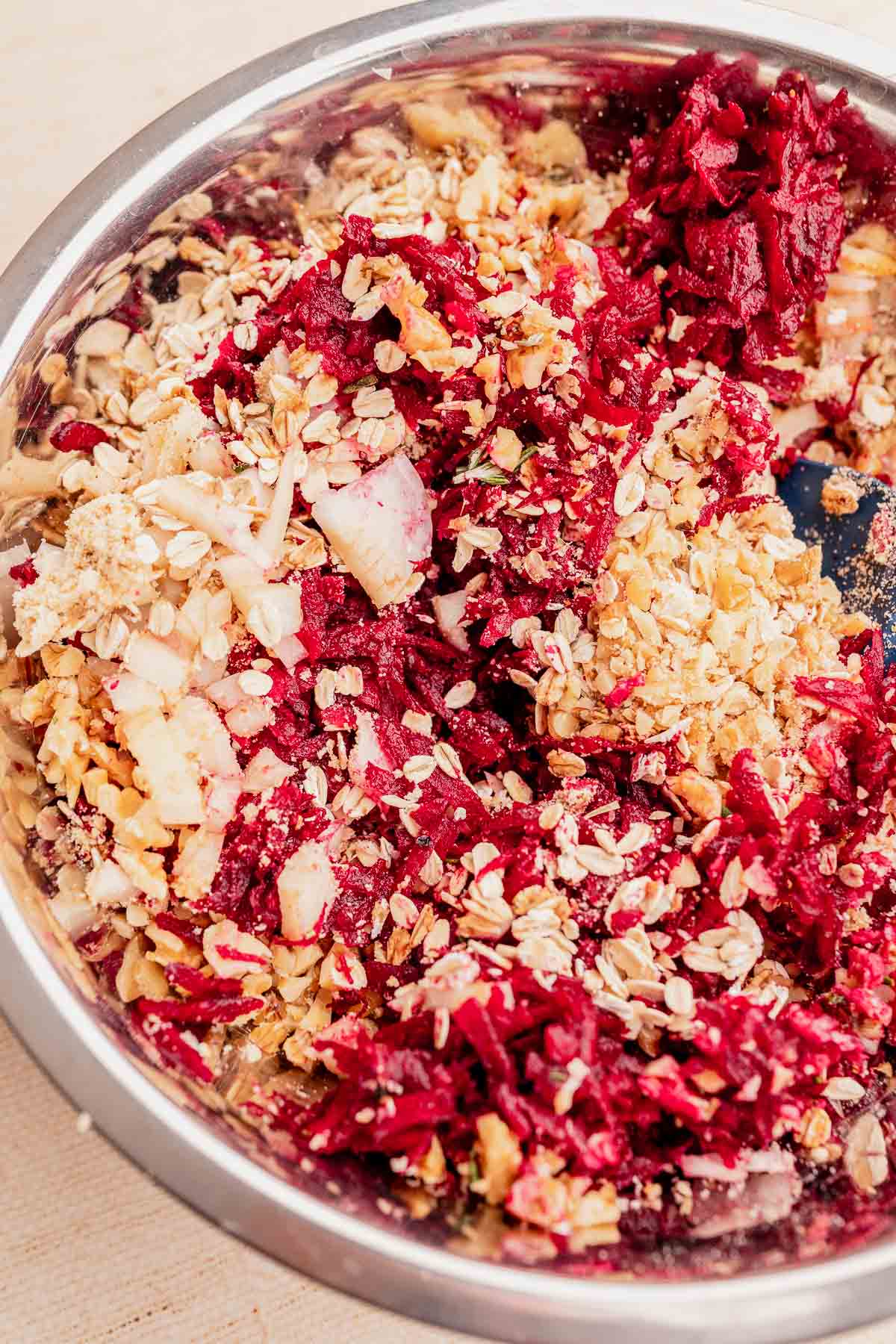 A stainless steel bowl contains a mixture of grated beets, oats, nuts, and shredded coconut—perfect for crafting a hearty vegetarian roast.