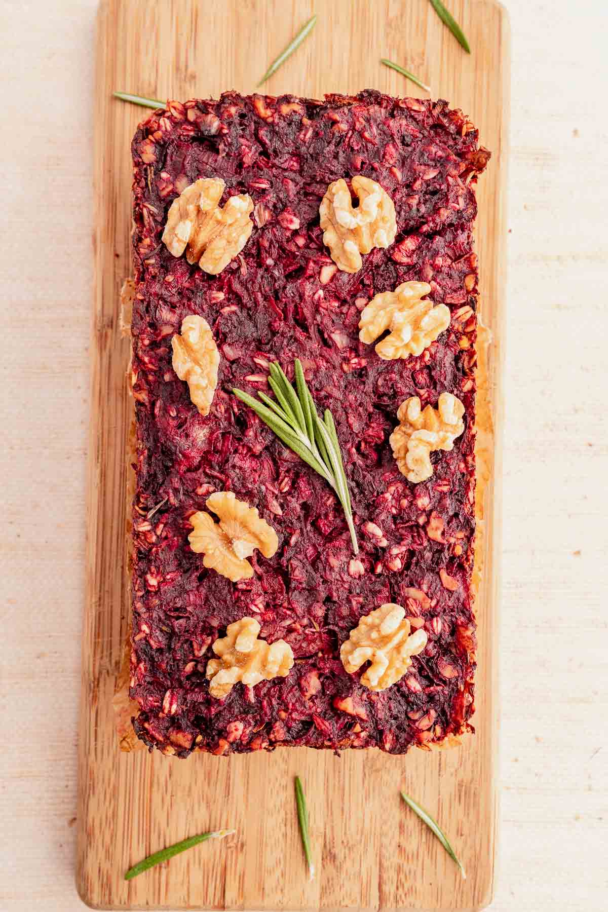 Loaf of beet and oat bread topped with walnuts and a sprig of rosemary, resembling a savory vegetarian roast, placed on a wooden board.