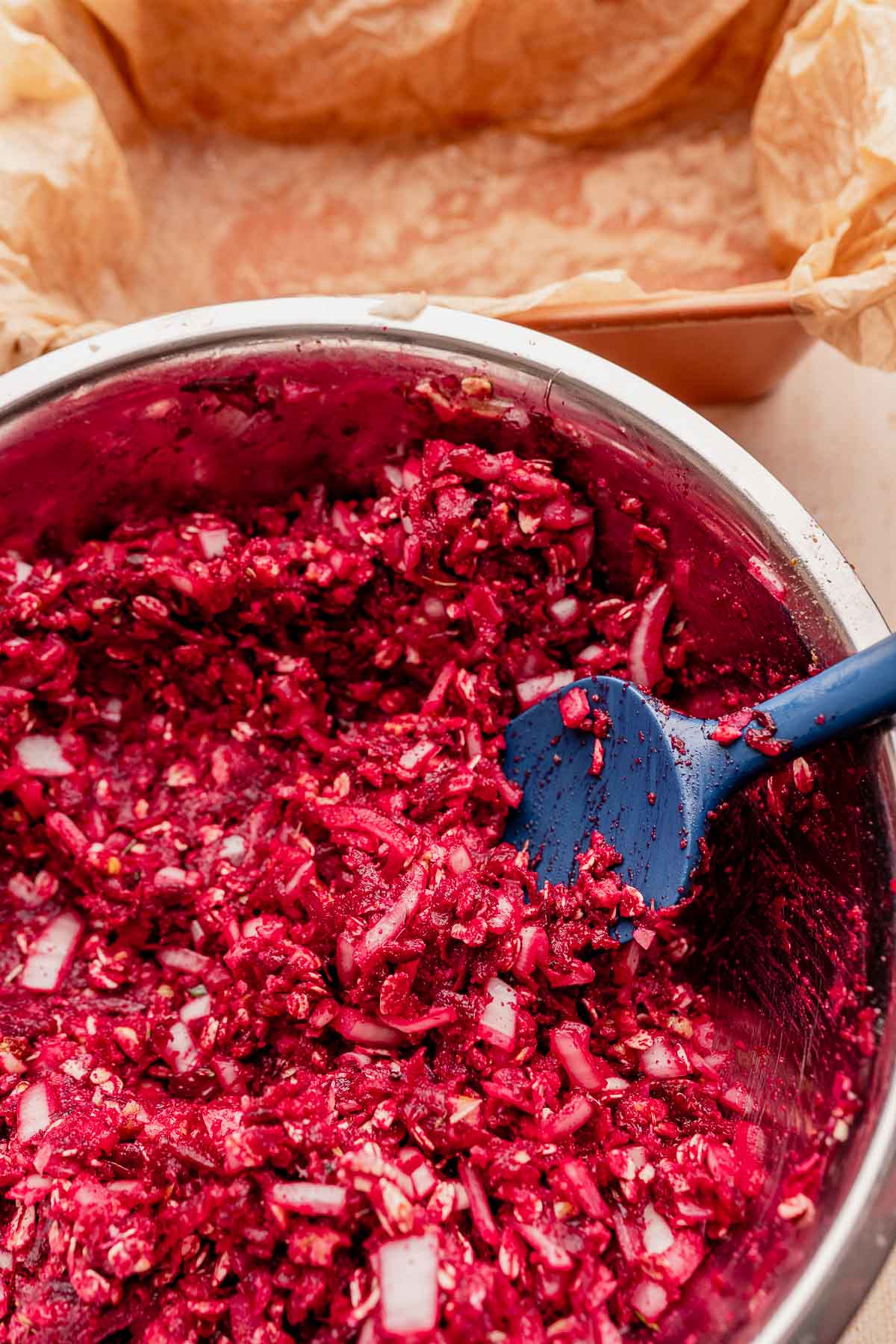 A bowl of beetroot and diced onion mixture, perfect for crafting a delicious vegetarian roast, sits alongside a blue spatula next to a parchment-lined baking pan.