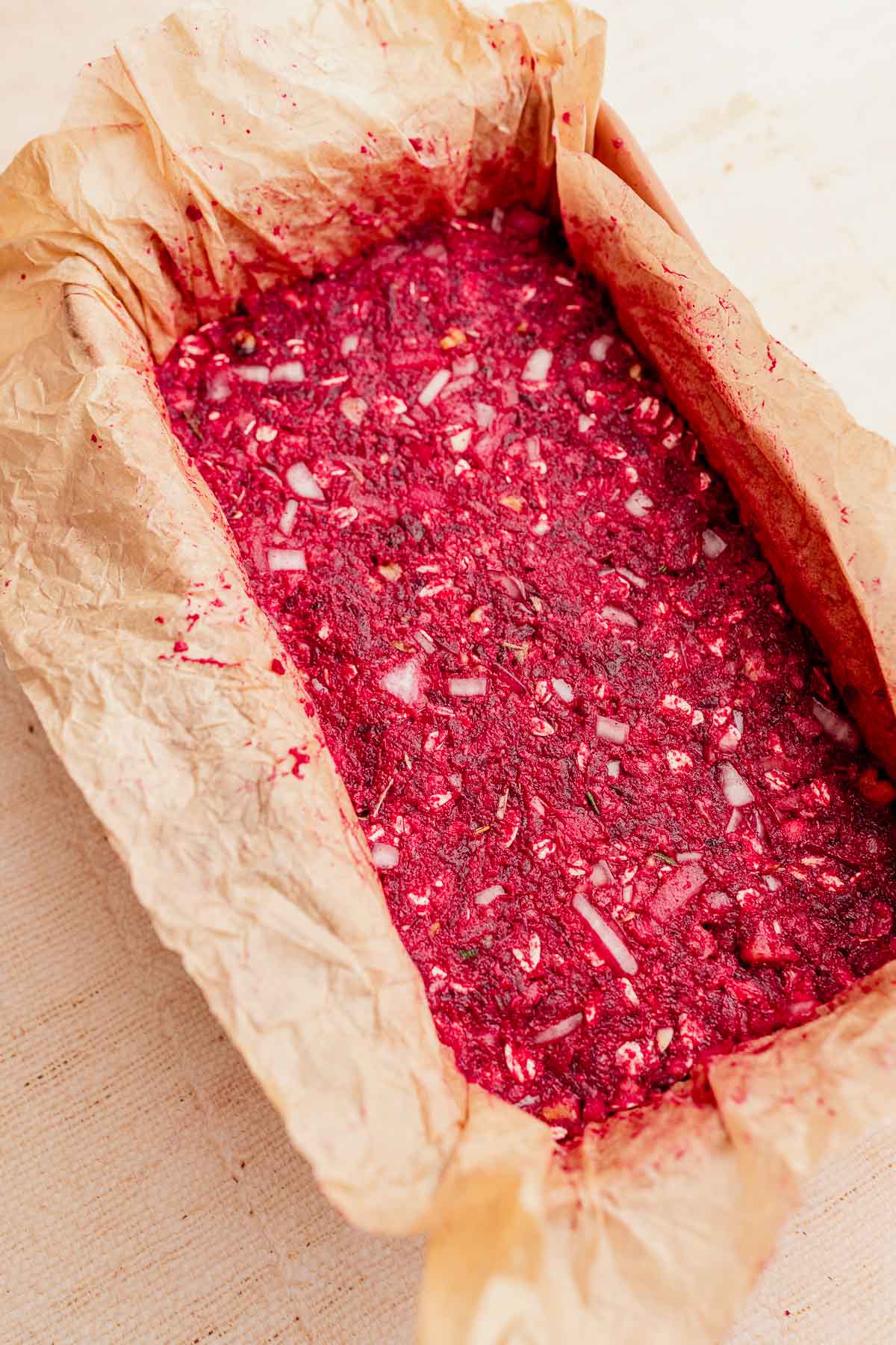 A rectangular baking dish lined with parchment paper contains a vibrant vegetarian roast, featuring a red beet mixture adorned with visible onions and fragrant herbs.