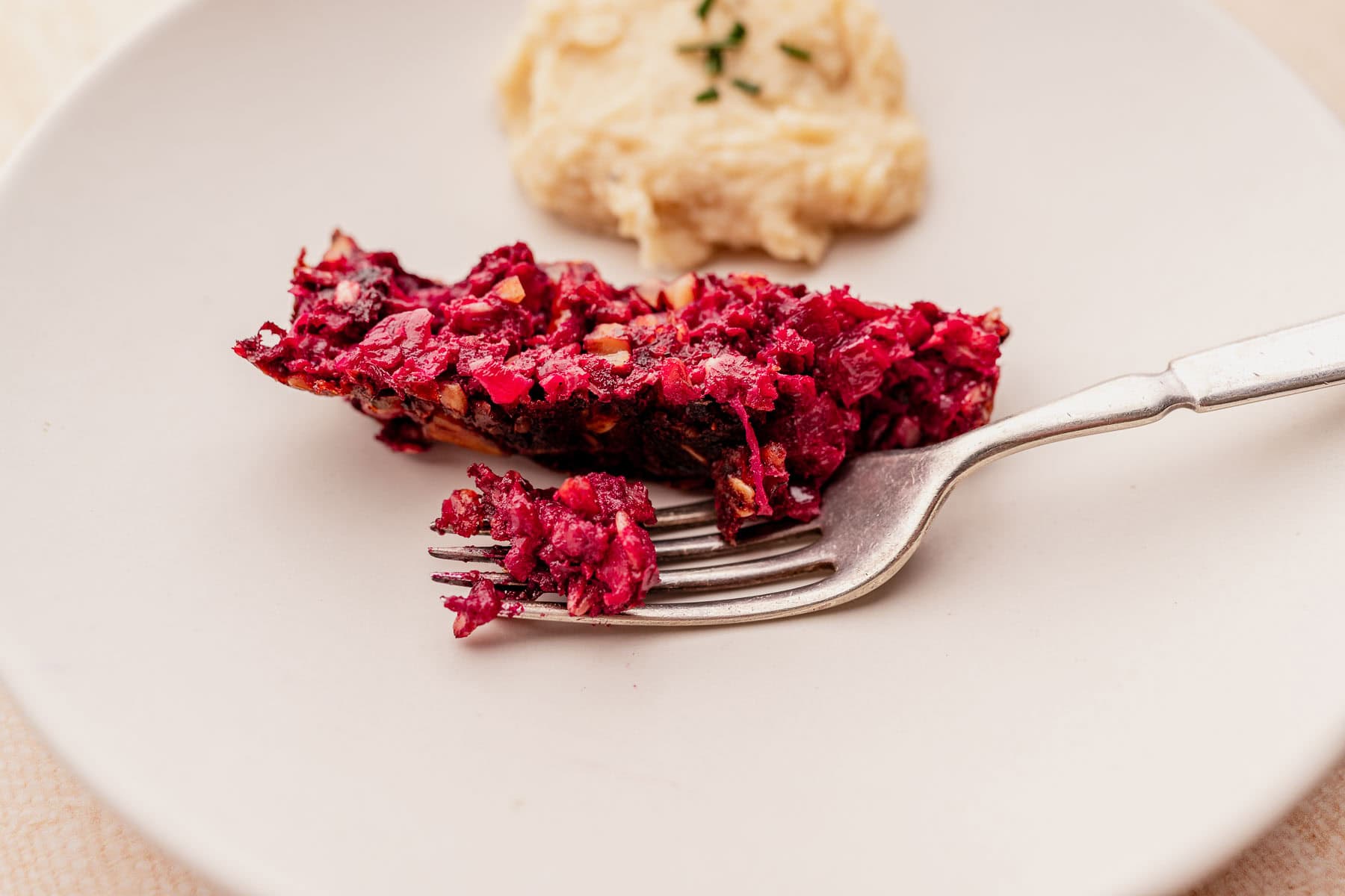 A fork holding a piece of beetroot tart on a white plate, reminiscent of a vegetarian roast, with a dollop of beige pureed sauce in the background.