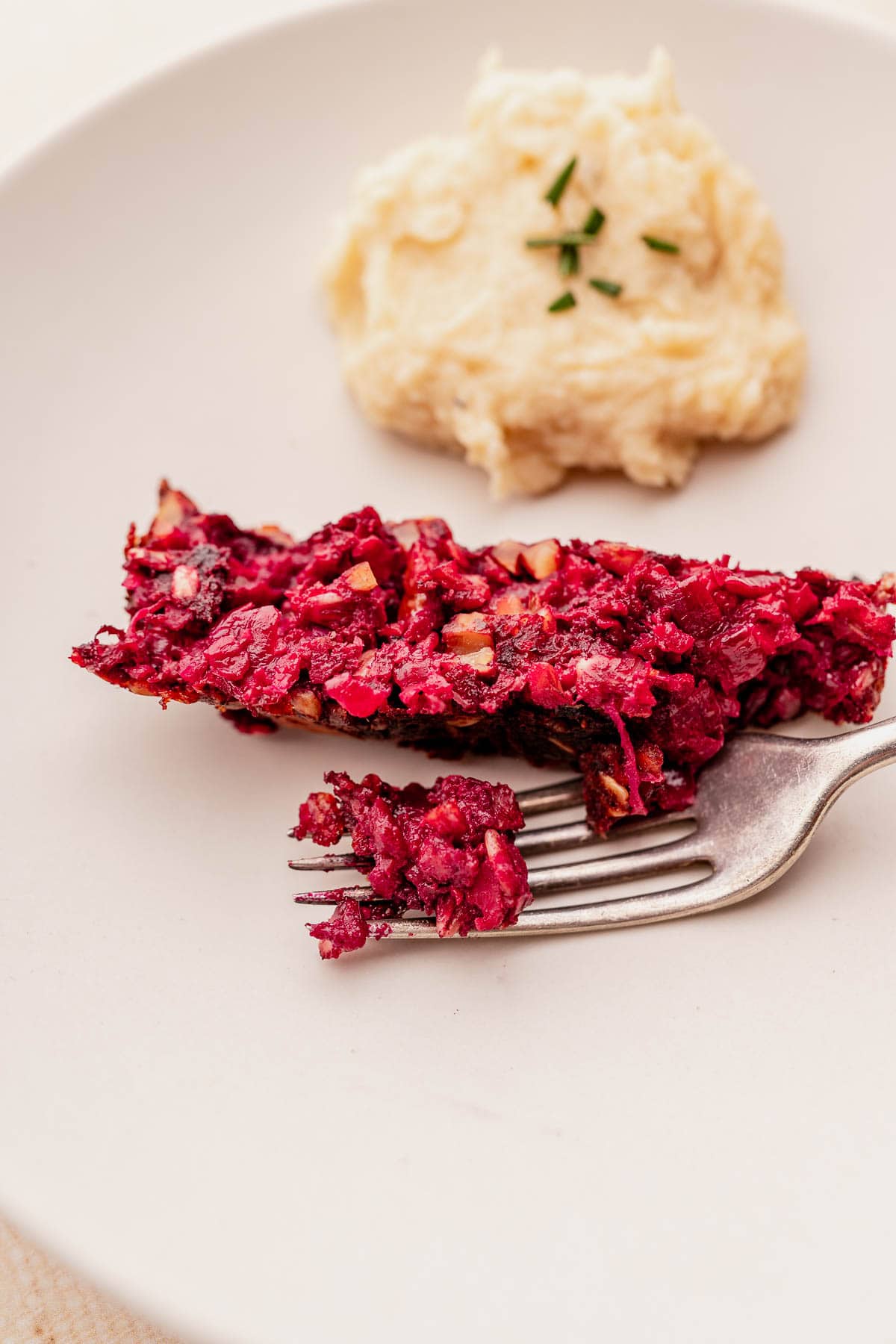 A slice of beetroot loaf on a plate with a fork, served alongside creamy mashed potatoes garnished with chives, makes for a delightful vegetarian roast experience.