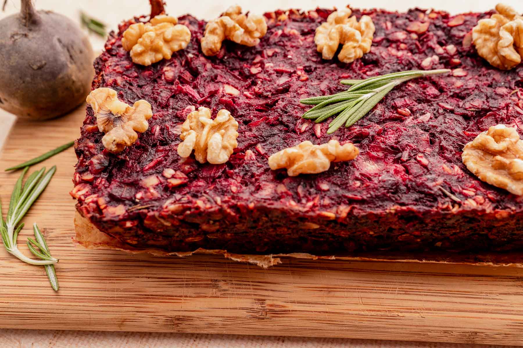 Close-up of a vegetarian roast beet loaf topped with walnuts and a sprig of rosemary on a wooden board, accompanied by additional rosemary on the side.