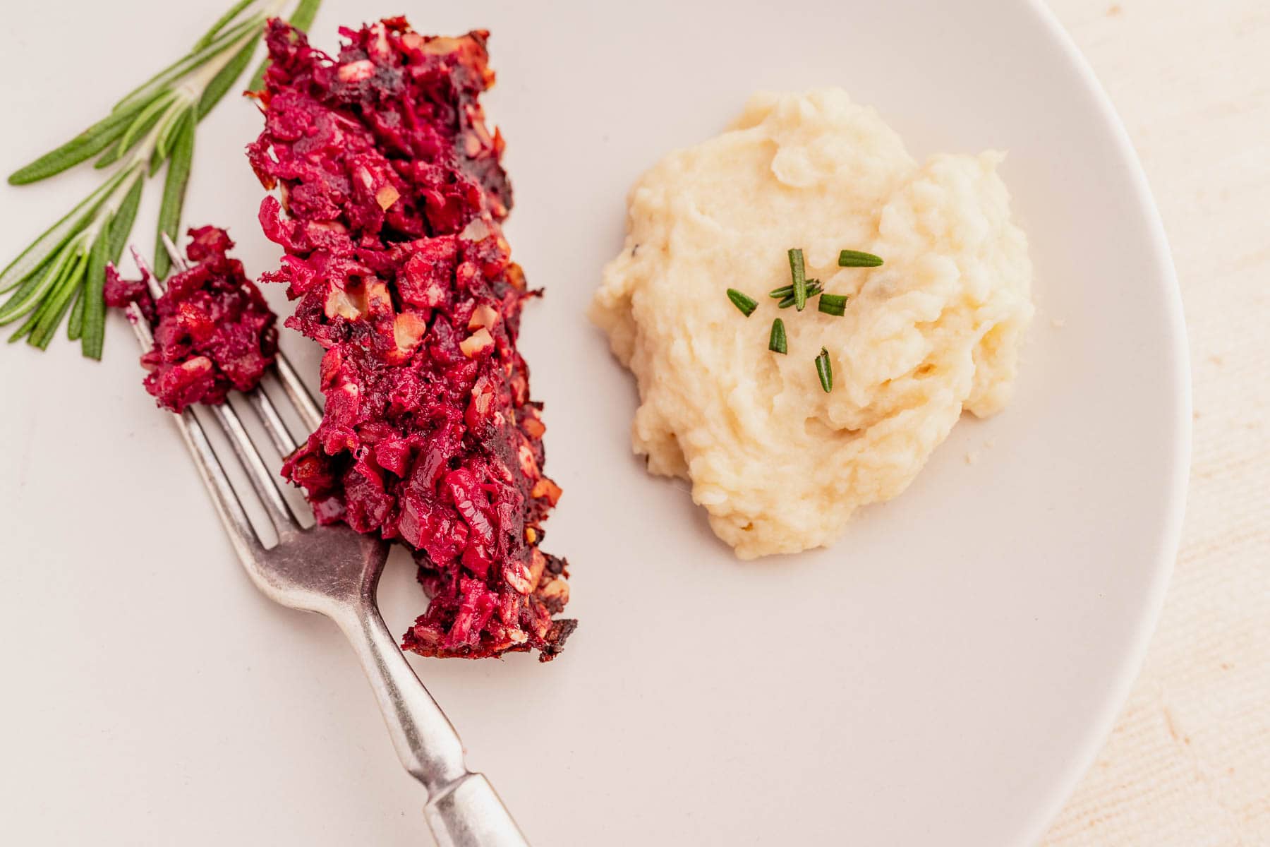 A plate with beetroot casserole and mashed potatoes, garnished with chives and sprigs of rosemary, evokes the essence of a delightful vegetarian roast, with a fork resting on the side.