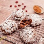 Chocolate cranberry crinkle cookies rest on a cooling rack, dusted with powdered sugar. One cookie is broken open to reveal its luscious filling. In the background, dried cranberries and a spoonful of cinnamon await their turn in holiday baking magic.