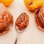 A spoonful of pear jam rests on a table beside glass jars of jam and two fresh pears.