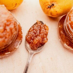 A spoonful of pear jam rests on a table beside glass jars of jam and two fresh pears.