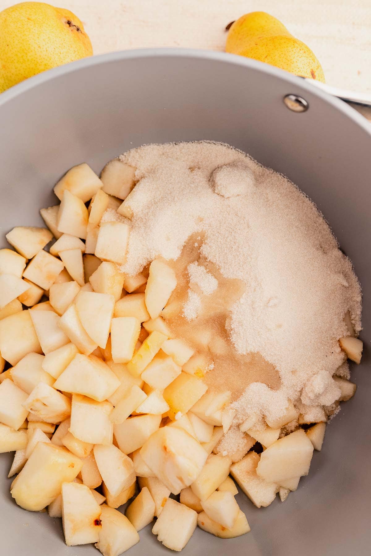 Chopped pears and sugar in a gray pot, with two whole pears in the background.