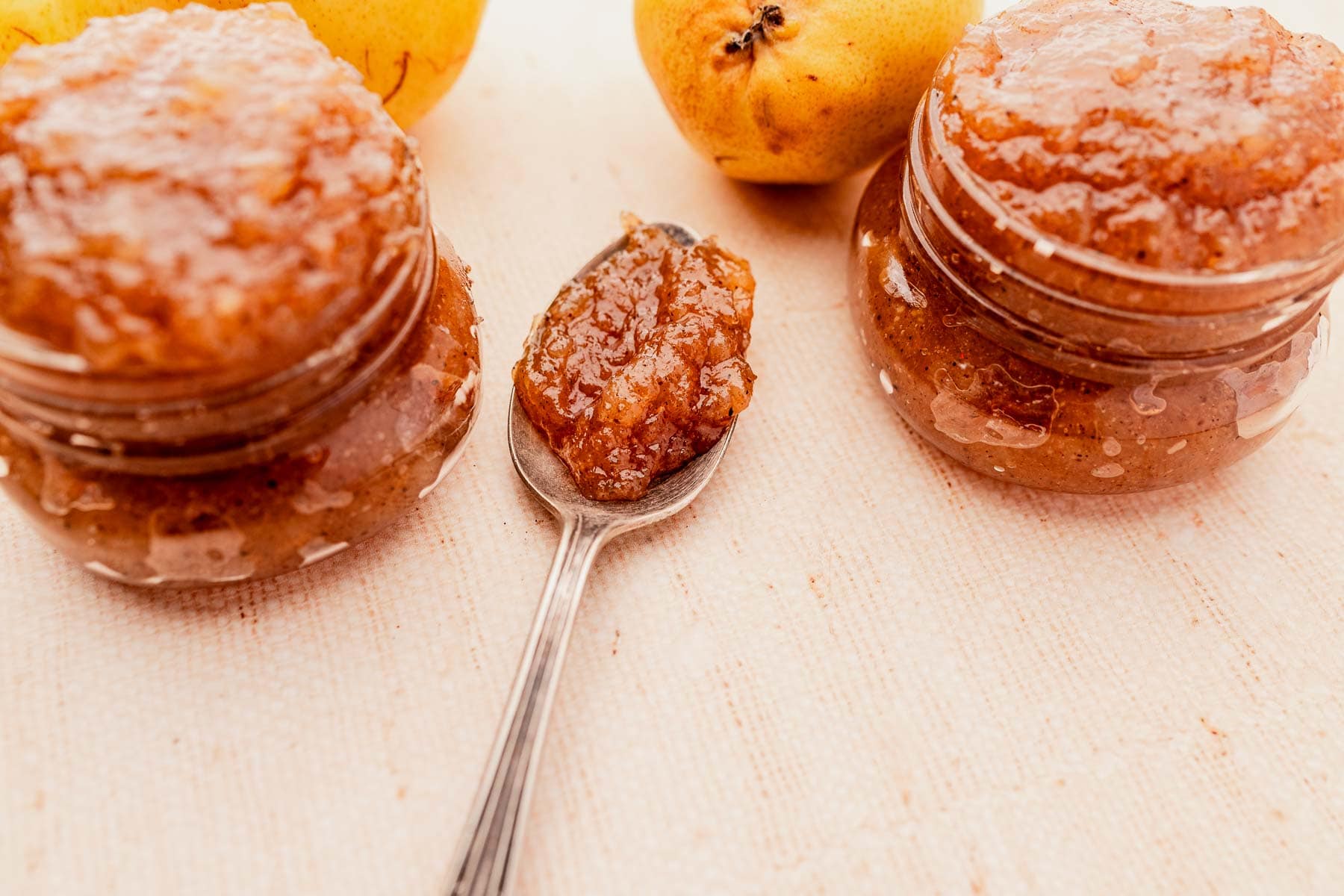 Jars of fruit jam and a spoonful of jam on a light cloth surface with two yellow fruits in the background.