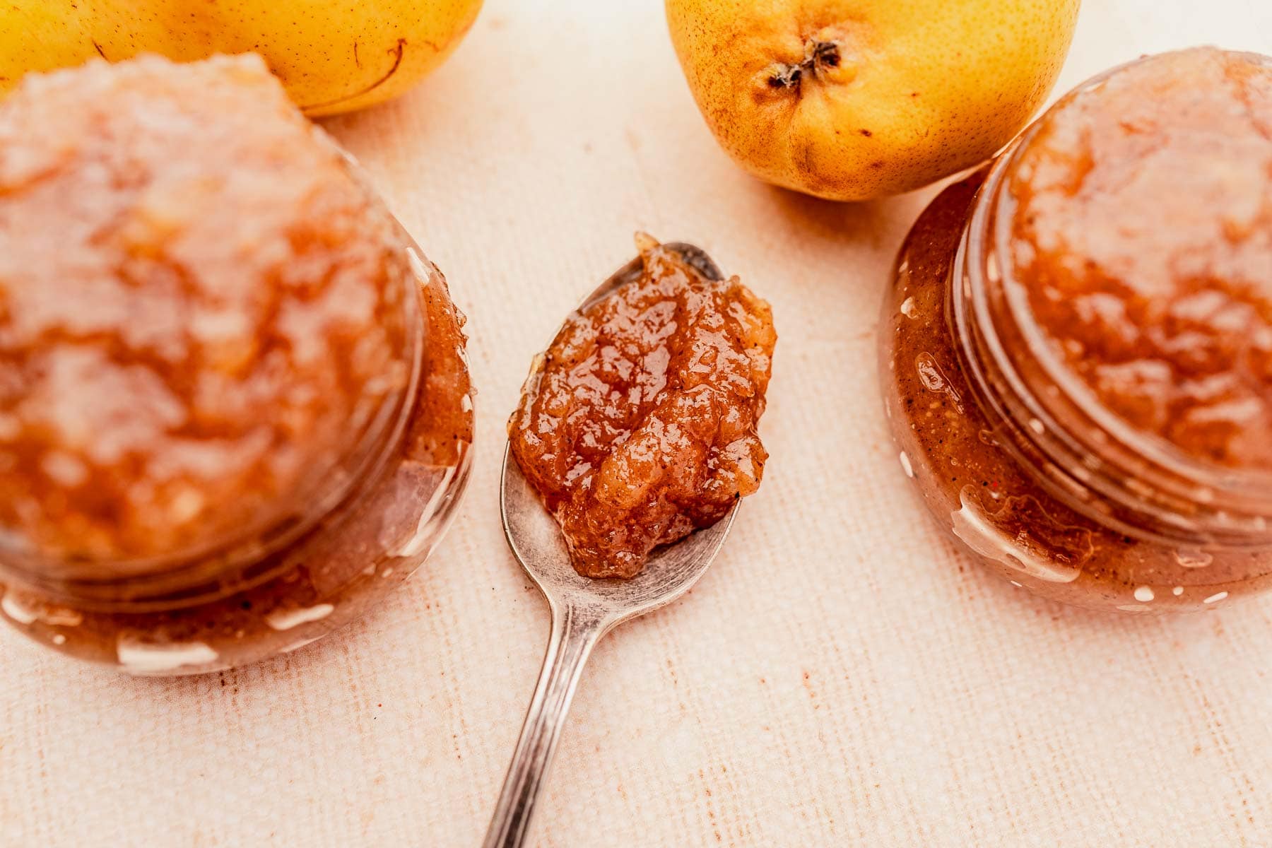 Jars of pear jam with a spoonful placed between them on a white surface, surrounded by fresh pears.