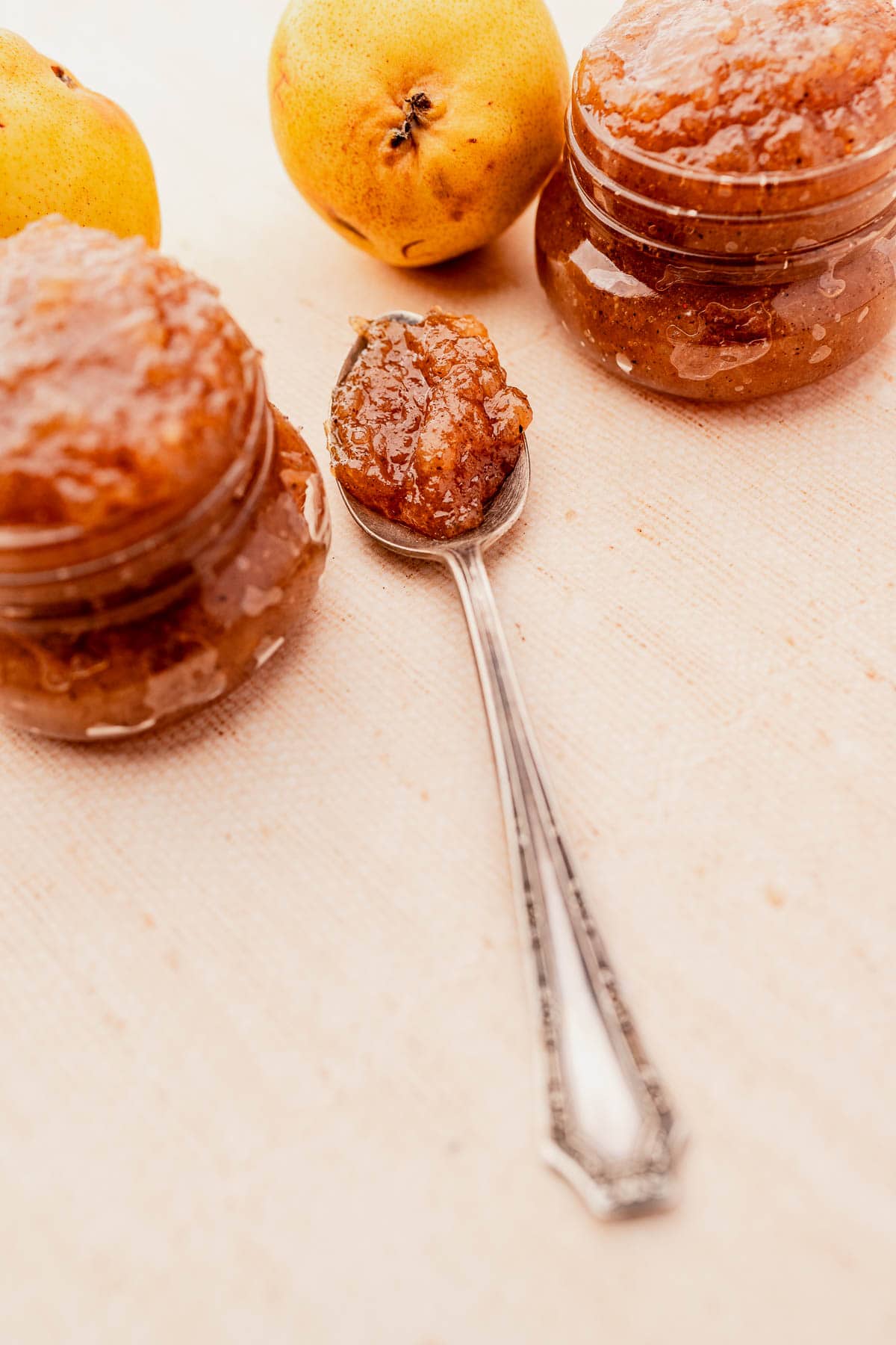 A spoon with fruit preserve sits on a light surface, surrounded by two small jars of the same preserve and a few whole pears.