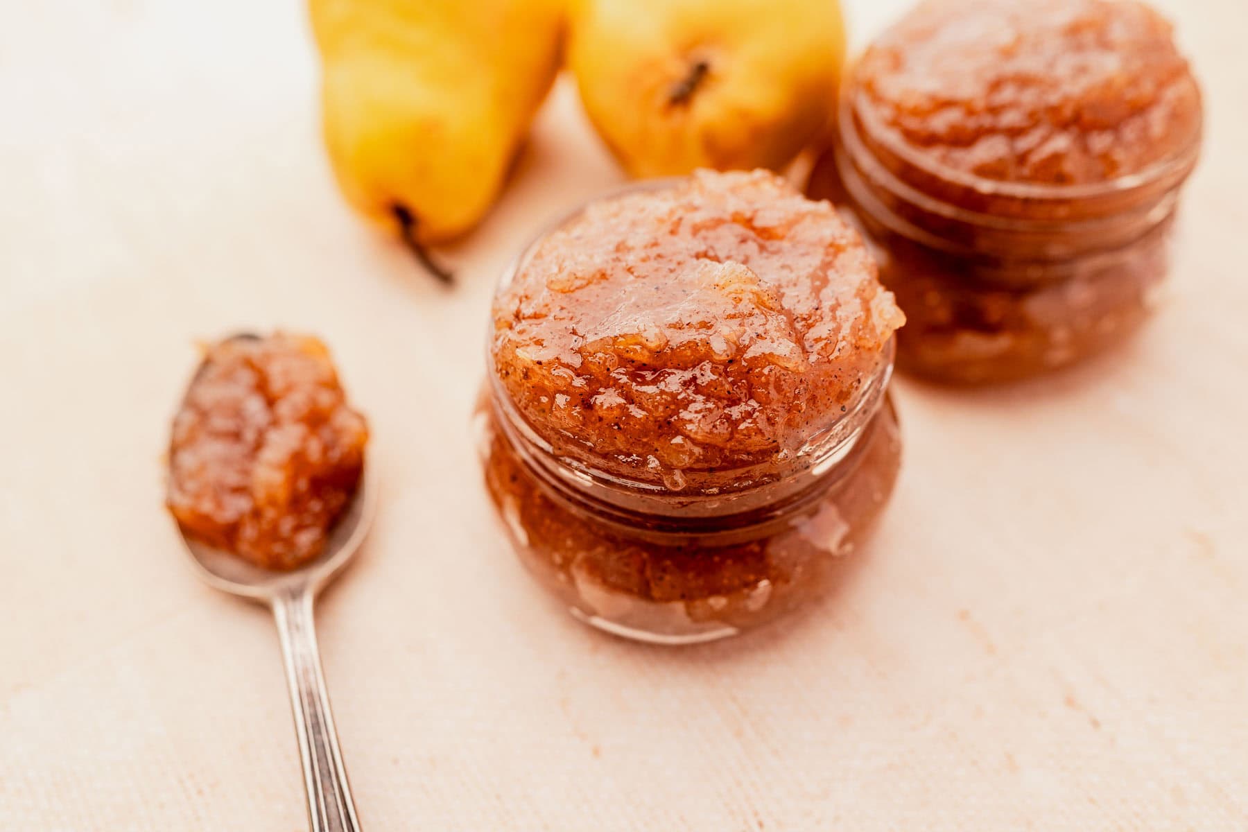 Two jars of pear jam and a spoonful of jam are placed on a tablecloth. Three fresh pears are in the background.
