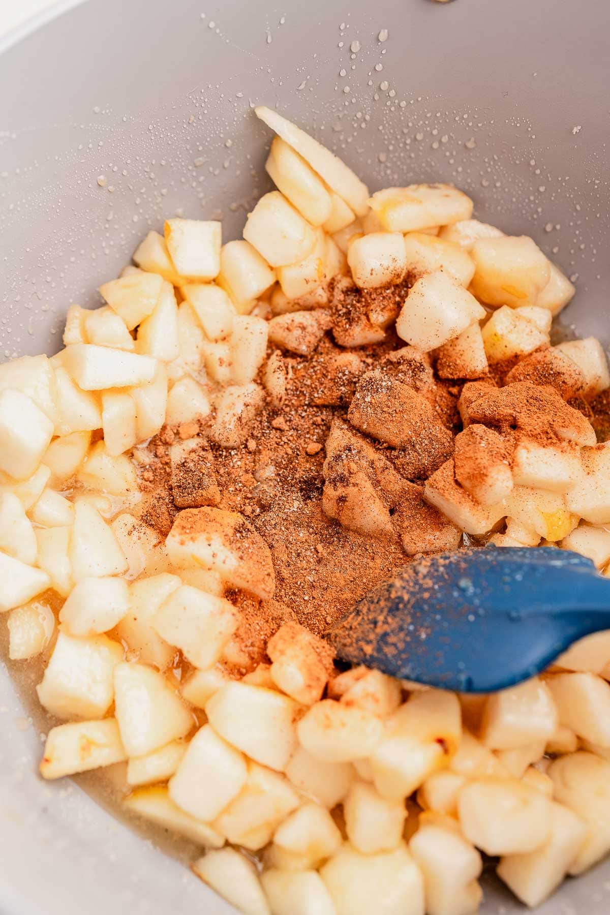 Diced apples mixed with cinnamon and spices in a bowl, with a blue spatula.