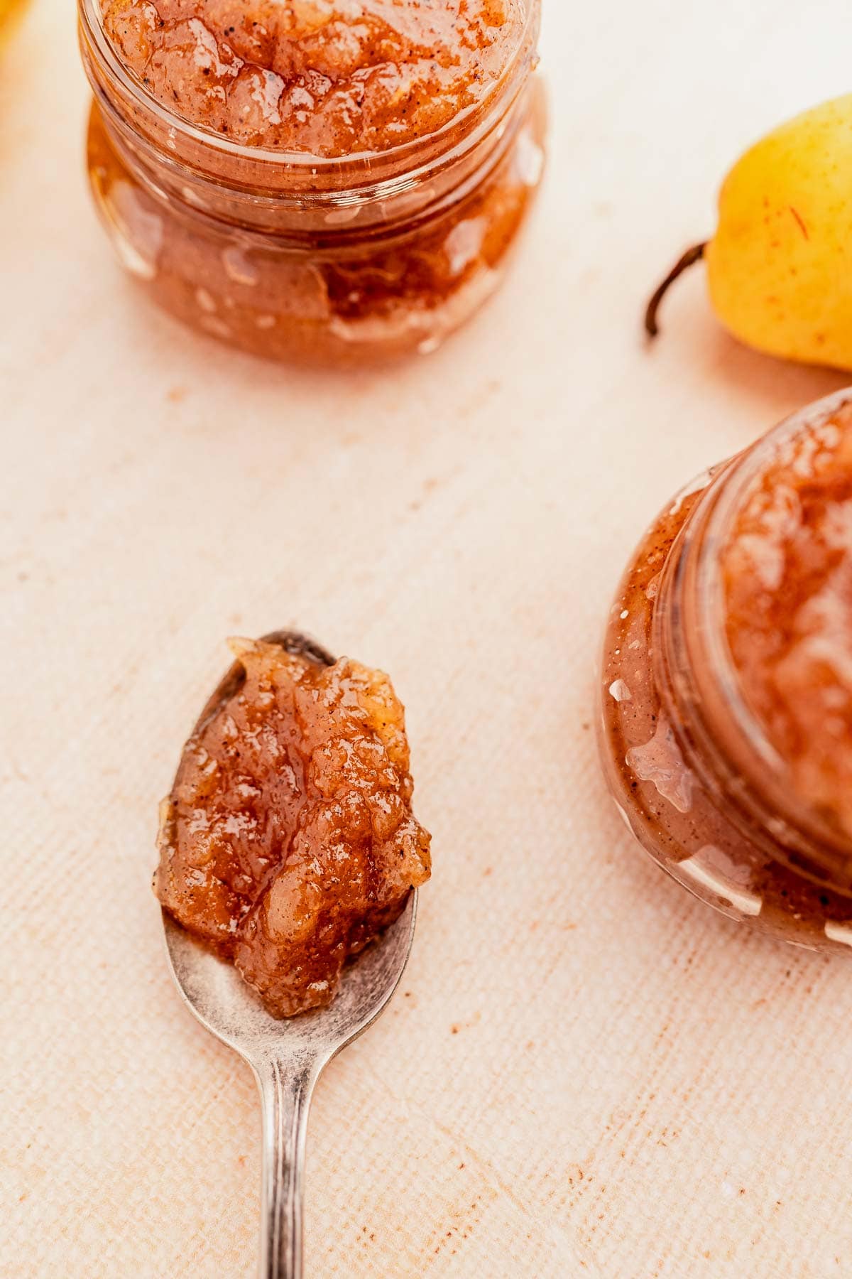 A spoonful of pear jam beside a jar of the same jam on a light surface, with a pear partially visible in the background.