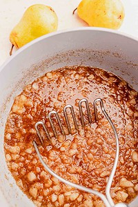 A metal potato masher mashes cooked pears in a pot, with two whole pears in the background.