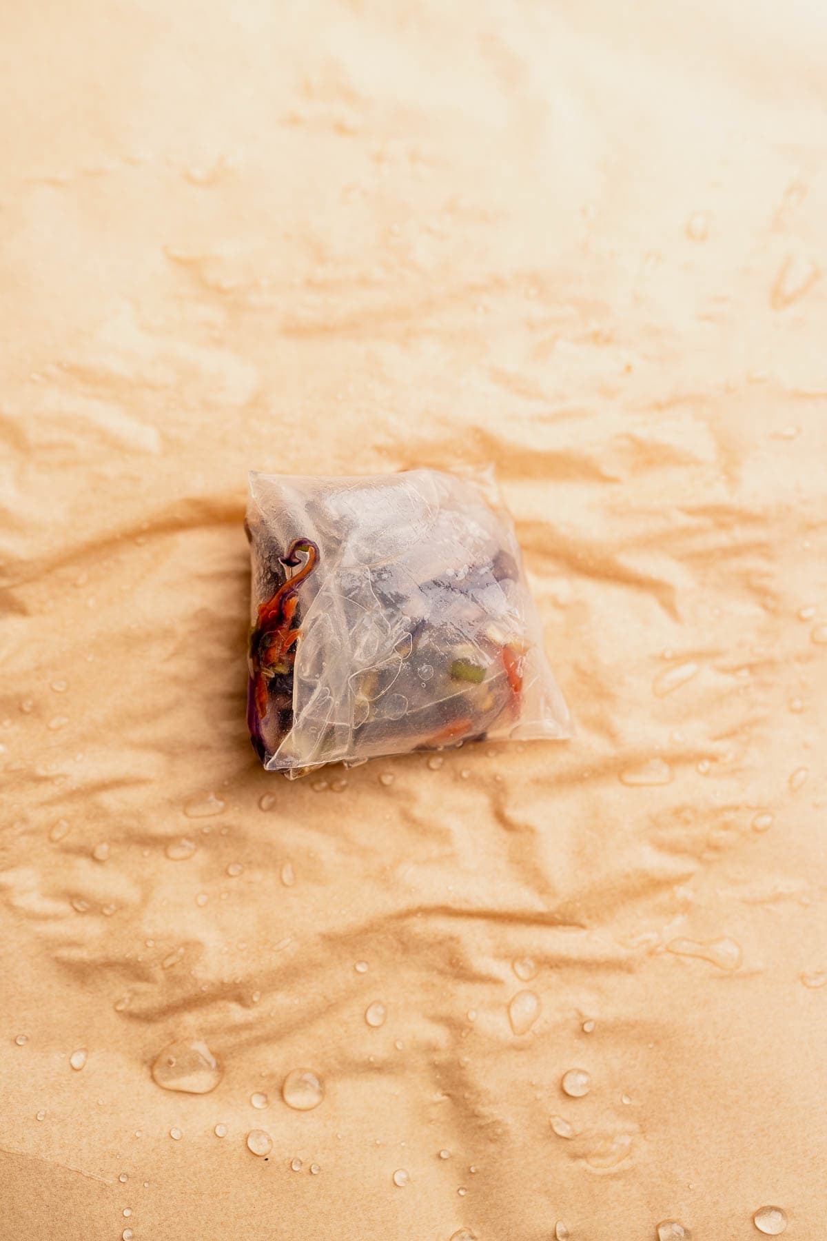 A sealed tea bag containing dried herbs and flowers rests on a textured, light brown surface with water droplets, reminiscent of the delicate beauty of air fryer rice paper dumplings.