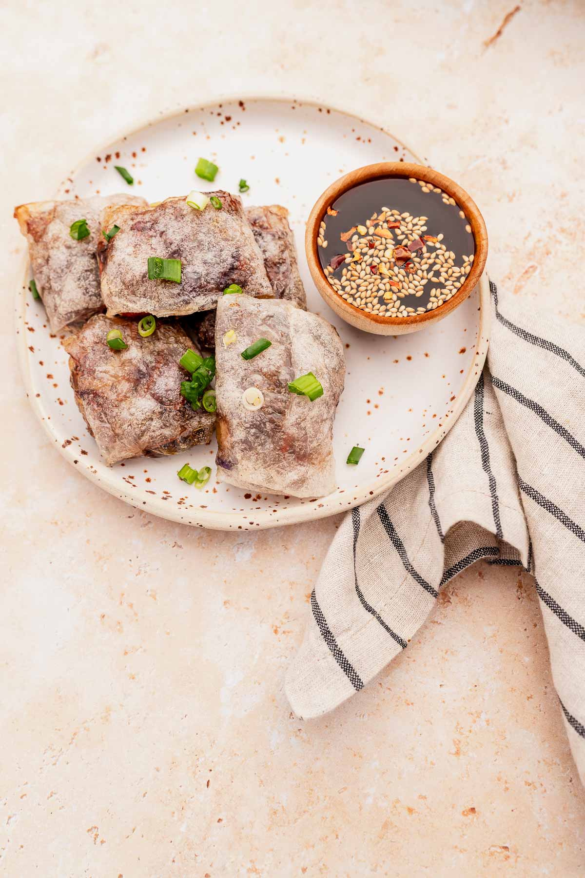 A plate with crispy air fryer dumplings garnished with green onions, accompanied by a bowl of dipping sauce with sesame seeds, and a striped napkin on the side.