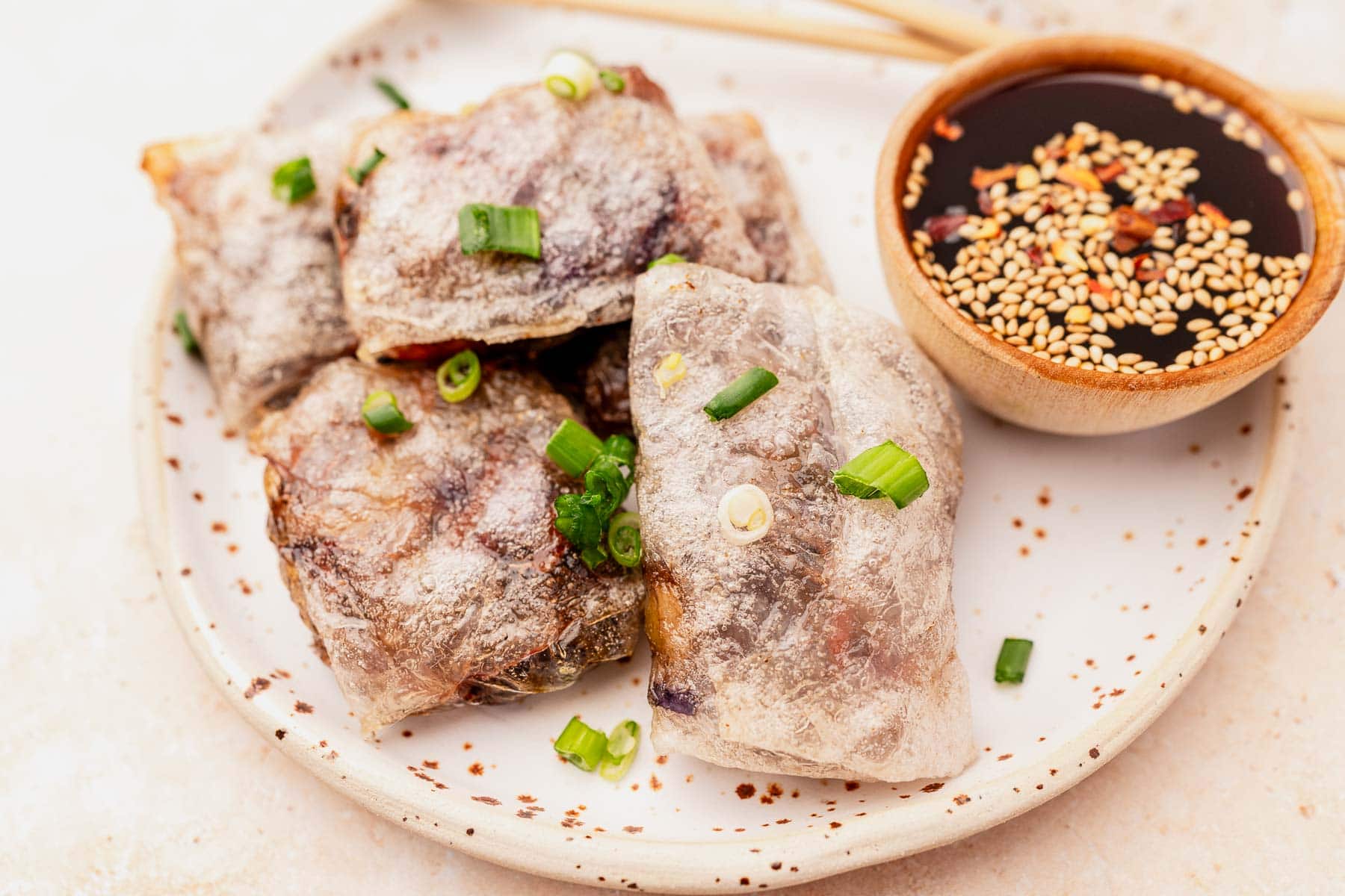 Four perfectly crisp air fryer rice paper dumplings with green onions rest on a plate next to a bowl of soy sauce sprinkled with sesame seeds.