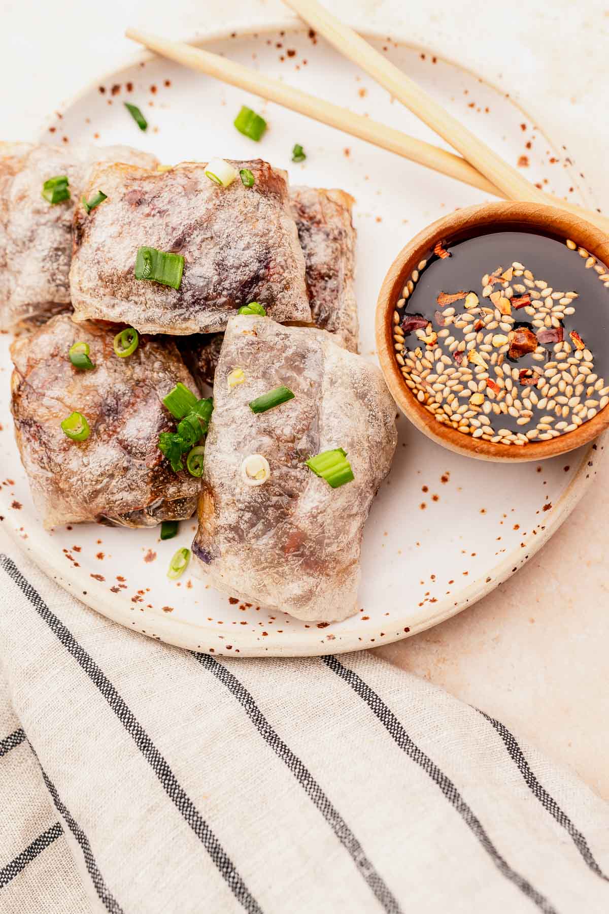A plate of translucent spring rolls, reminiscent of air fryer rice paper dumplings, is garnished with green onions and accompanied by a bowl of soy sauce sprinkled with sesame seeds and chili flakes. Chopsticks rest beside the plate on a striped cloth.