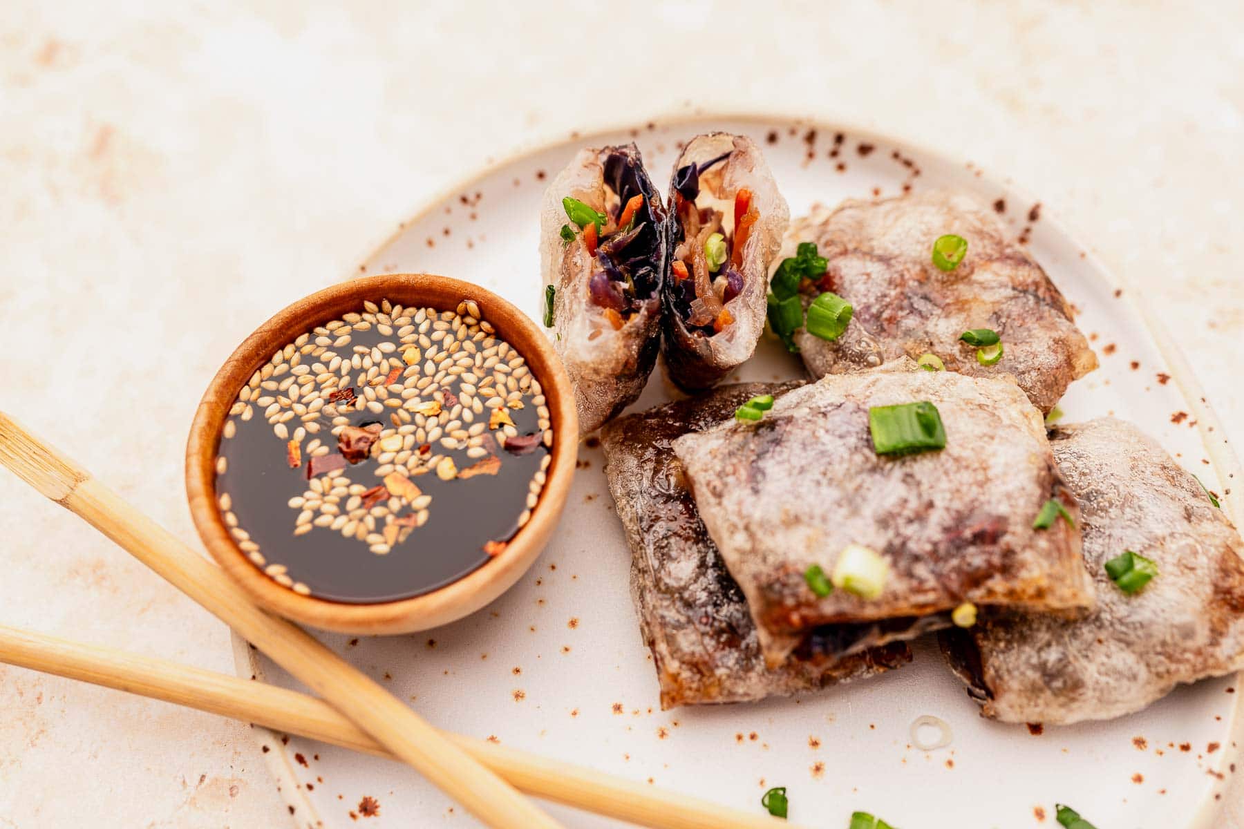 Plate with sliced crispy rolls filled with vegetables, garnished with green onions, next to a bowl of dipping sauce topped with sesame seeds. The rolls are air fryer rice paper dumplings, and chopsticks are on the side for easy eating.
