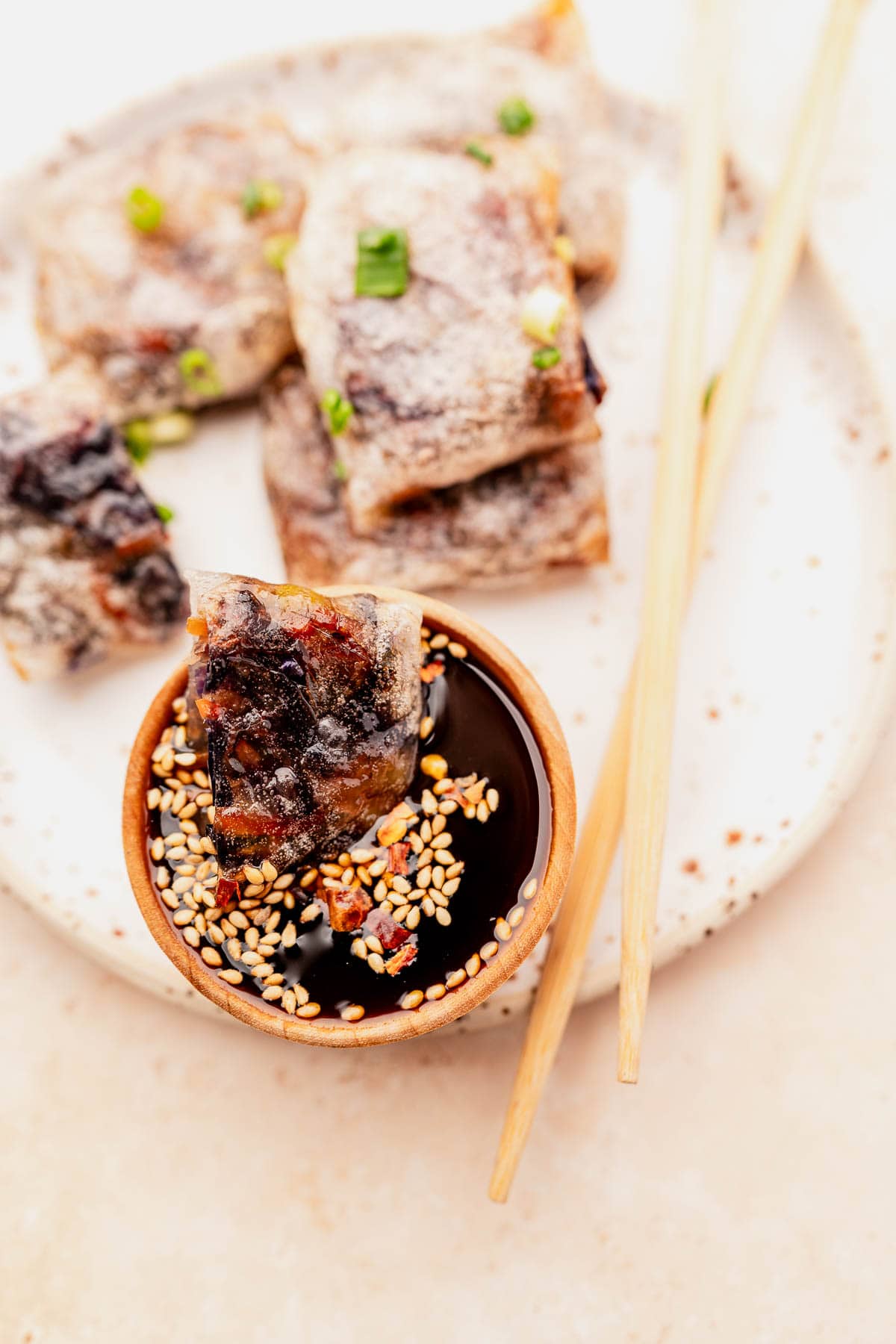 Crispy spring rolls on a plate with a bamboo bowl of soy sauce topped with sesame seeds, garnished with green onions. Perfectly cooked air fryer rice paper dumplings and chopsticks complete this delightful ensemble.
