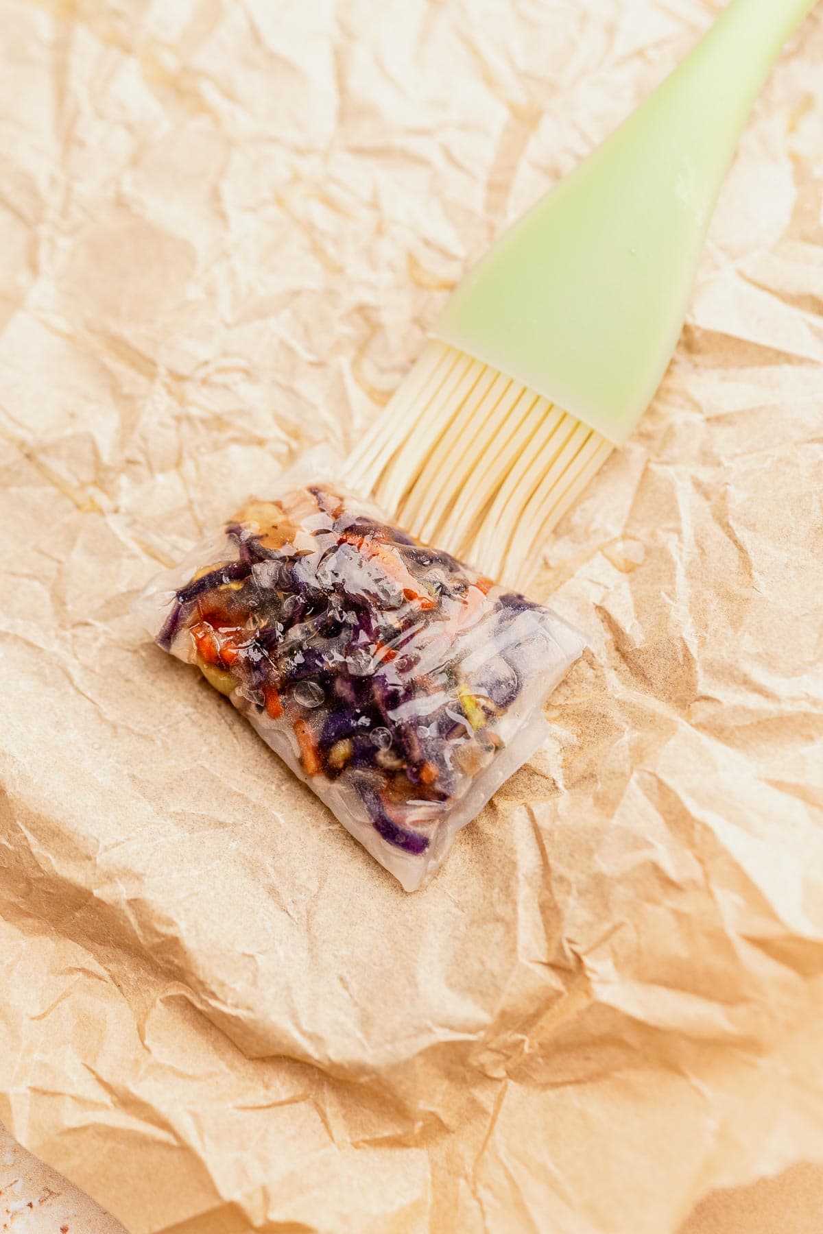 A rice paper roll filled with vegetables is brushed with oil using a light green silicone brush, placed on crumpled parchment paper, and transformed into crispy perfection in the air fryer.