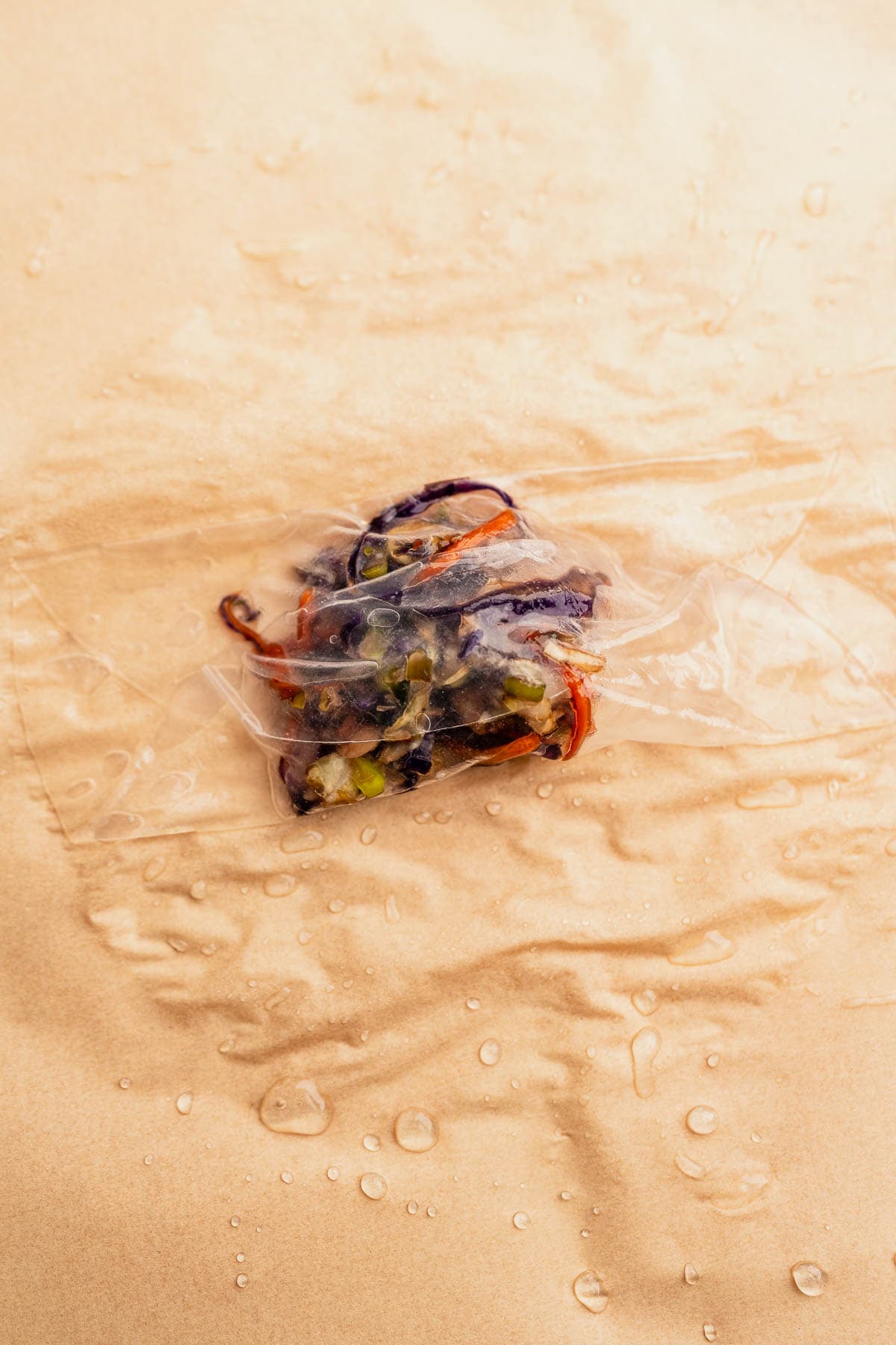A small bundle of colorful vegetable strips, perfect for stuffing air fryer rice paper dumplings, rests wrapped in clear plastic on a wet, light brown surface.