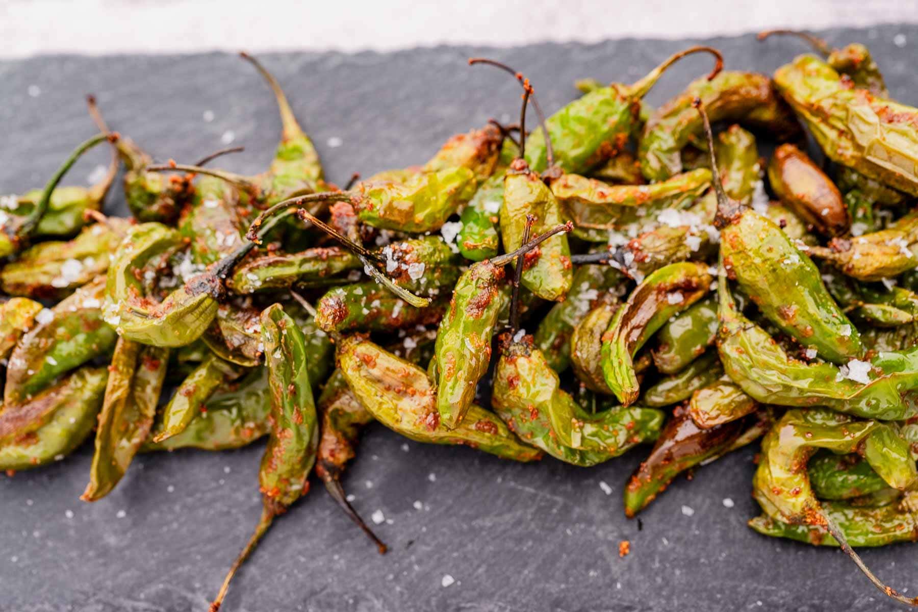 A pile of roasted shishito peppers, perfectly crispy from the air fryer and sprinkled with salt, rests on a dark slate surface.