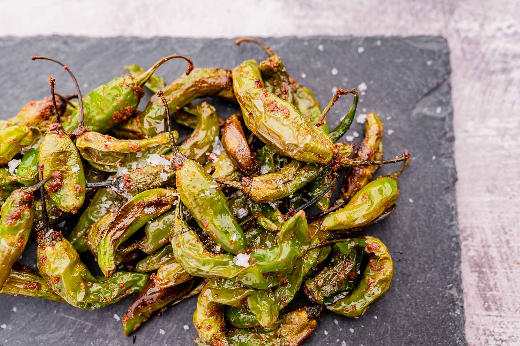 A pile of blistered Padron peppers sprinkled with coarse salt joins air fryer shishito peppers on a dark slate surface.